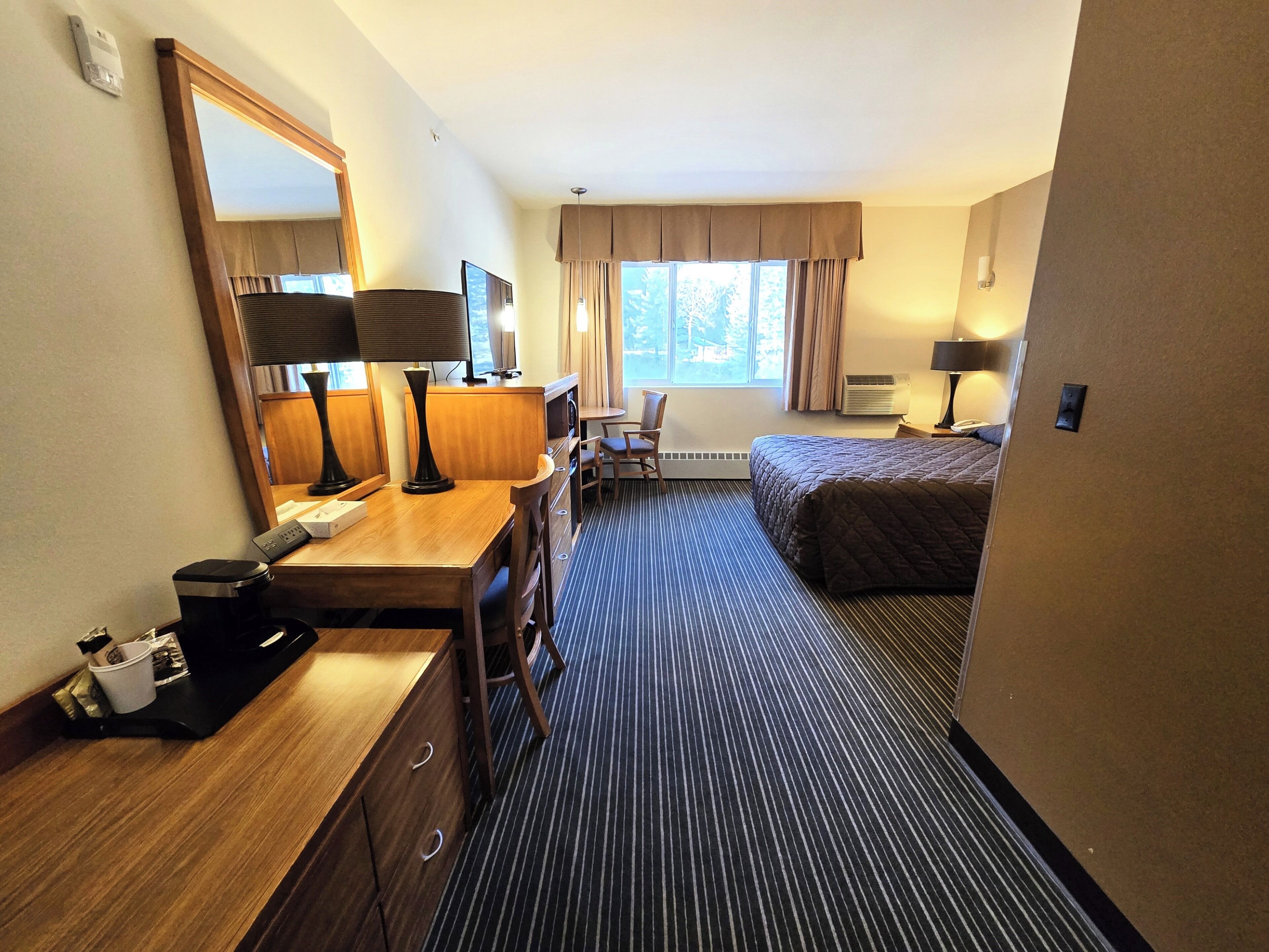 A hotel room with a bed, a desk with a chair, a mirror, a window with curtains, and a carpeted floor.