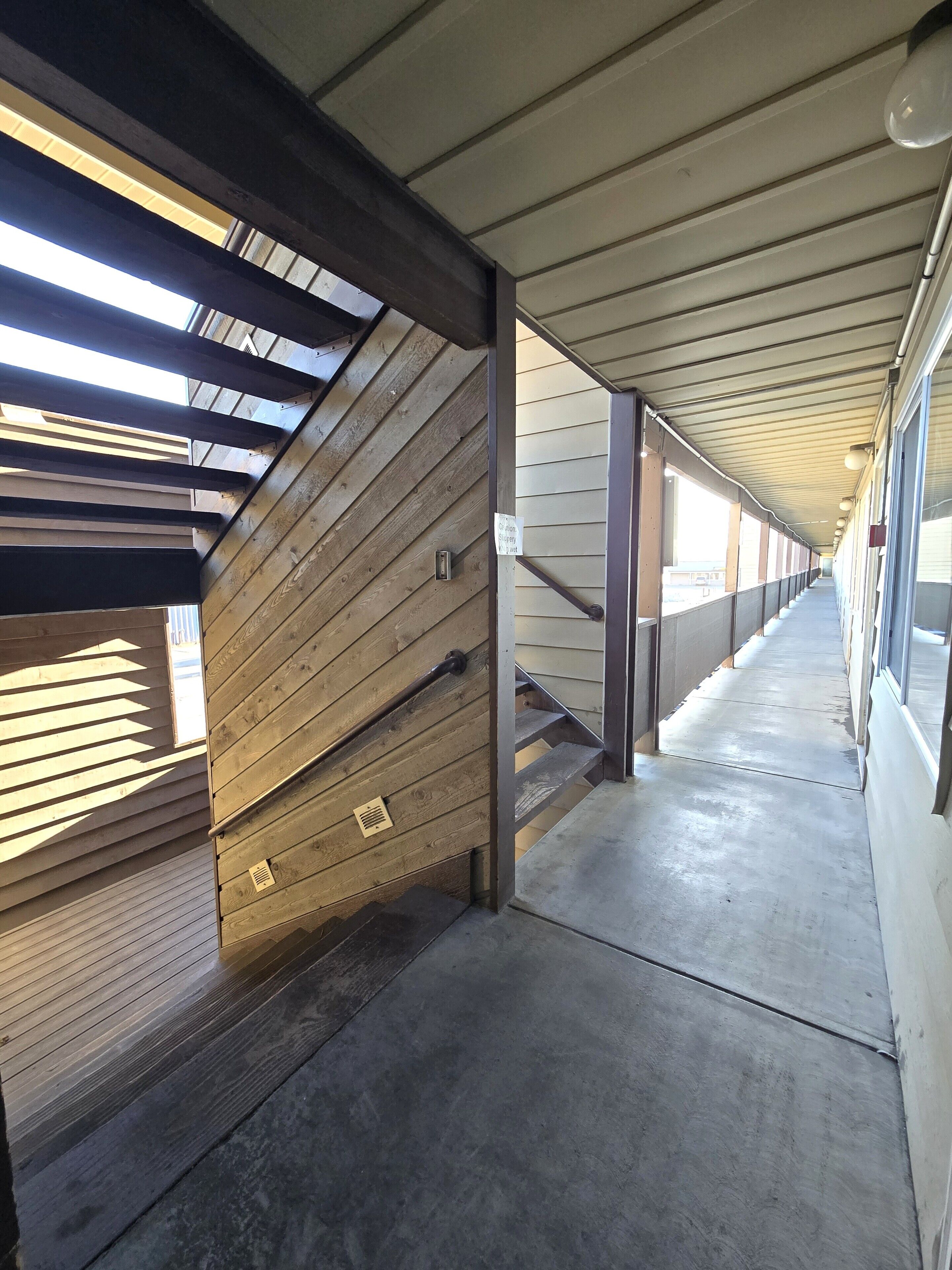 Un couloir couvert, avec des lambris en bois et des poutres métalliques, qui mène à un escalier.