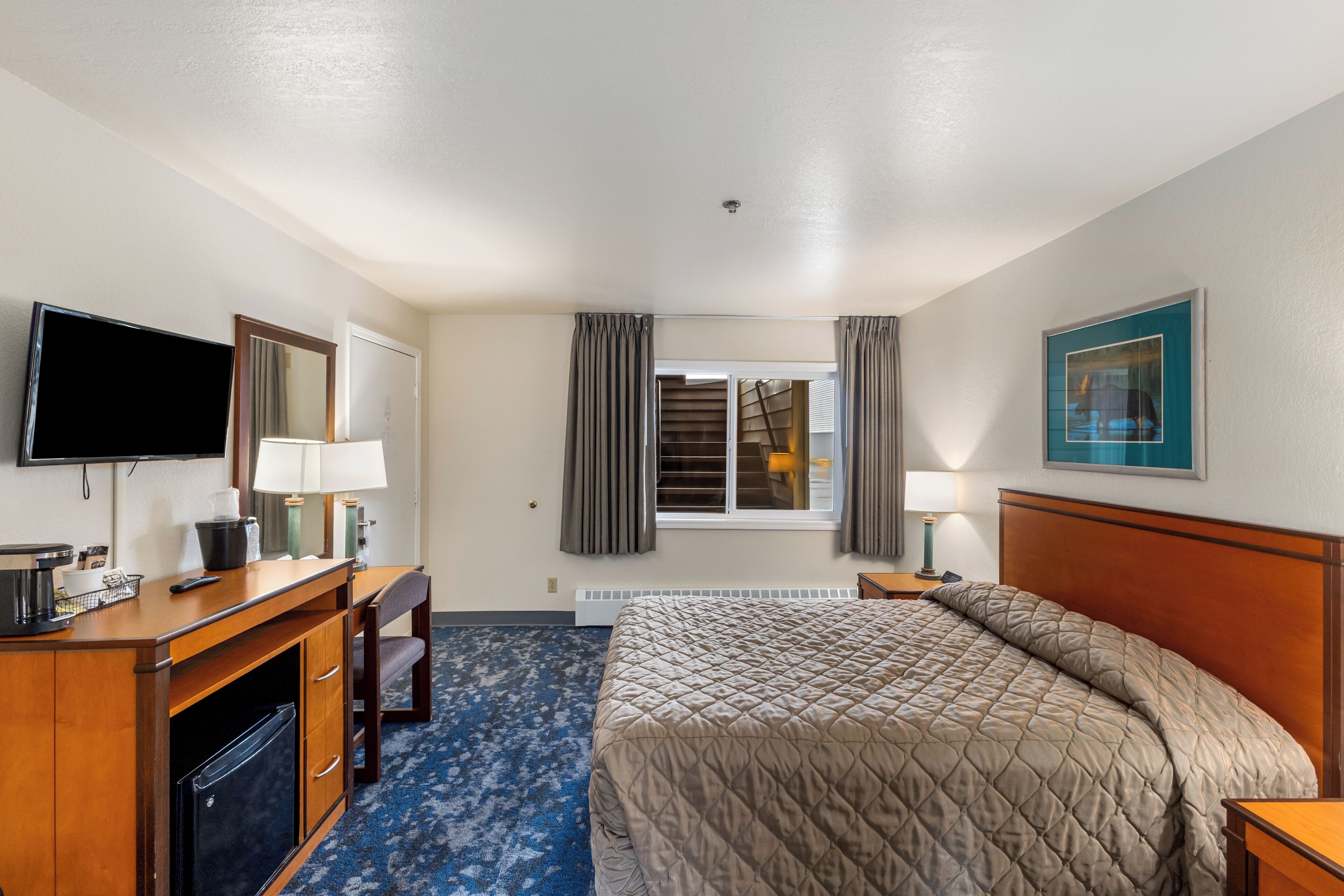 A hotel room with a bed, a desk with a TV, a chair, a lamp, and a window with curtains.