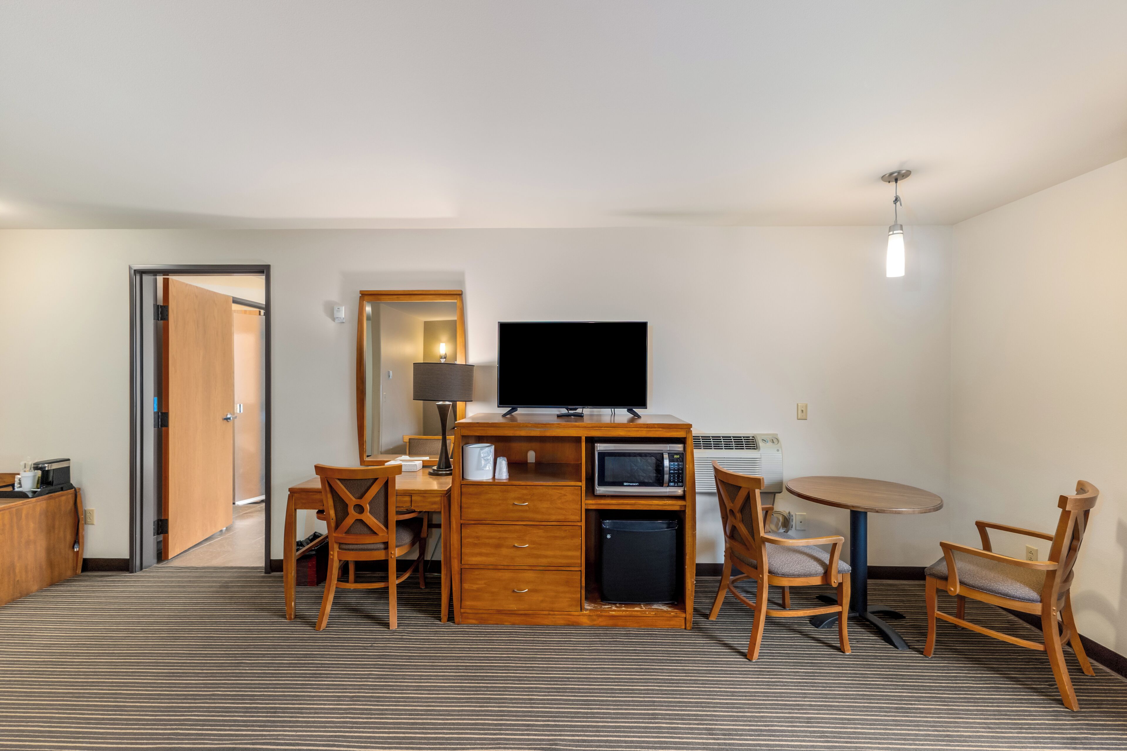 A hotel room with a desk, a television, a microwave, a refrigerator, a coffee maker, and a dining area with chairs.