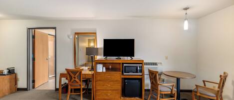 A hotel room with a desk, a television, a microwave, a refrigerator, a coffee maker, and a dining area with chairs.