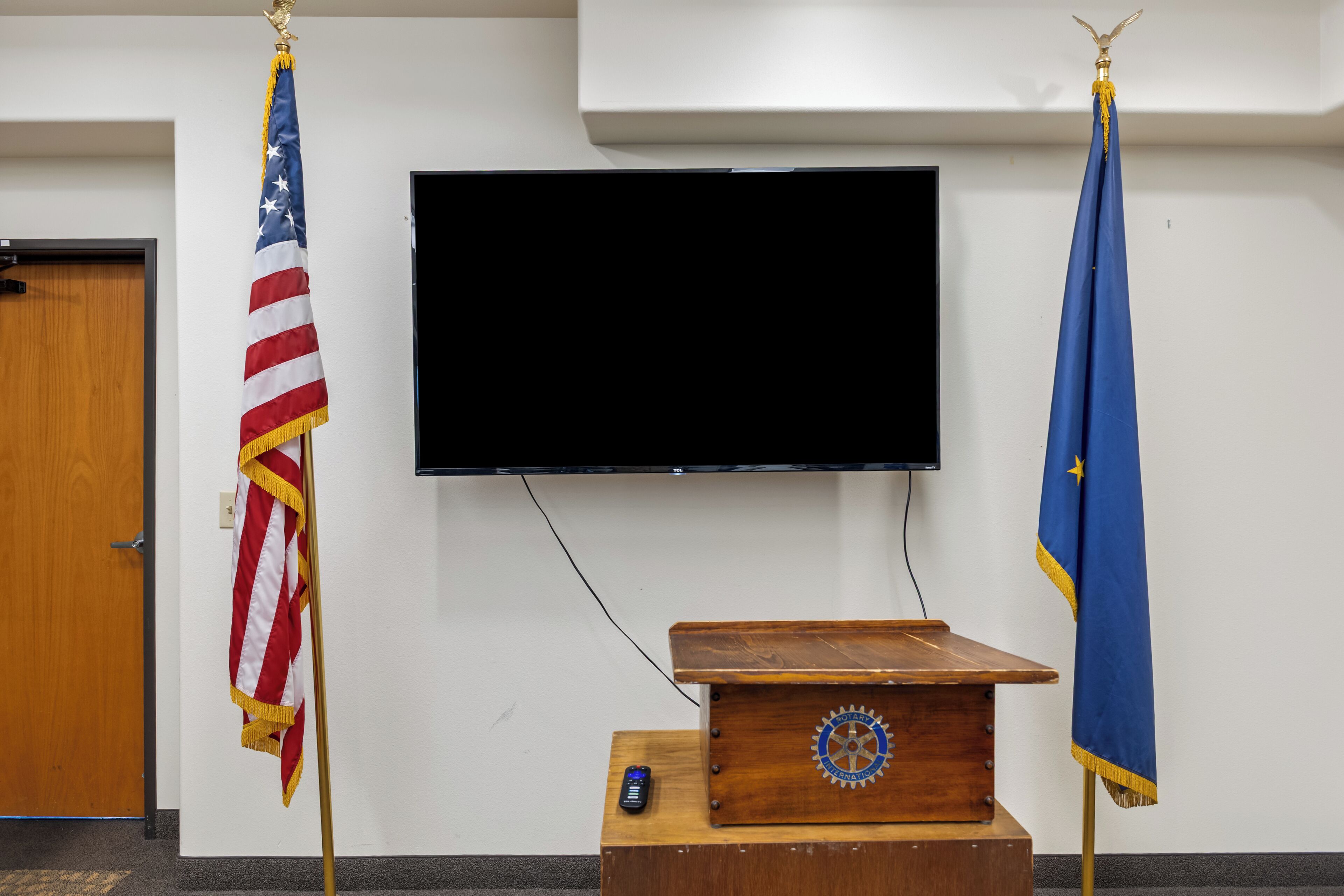 A room with a wooden podium, a television mounted on the wall, and two flags on stands.