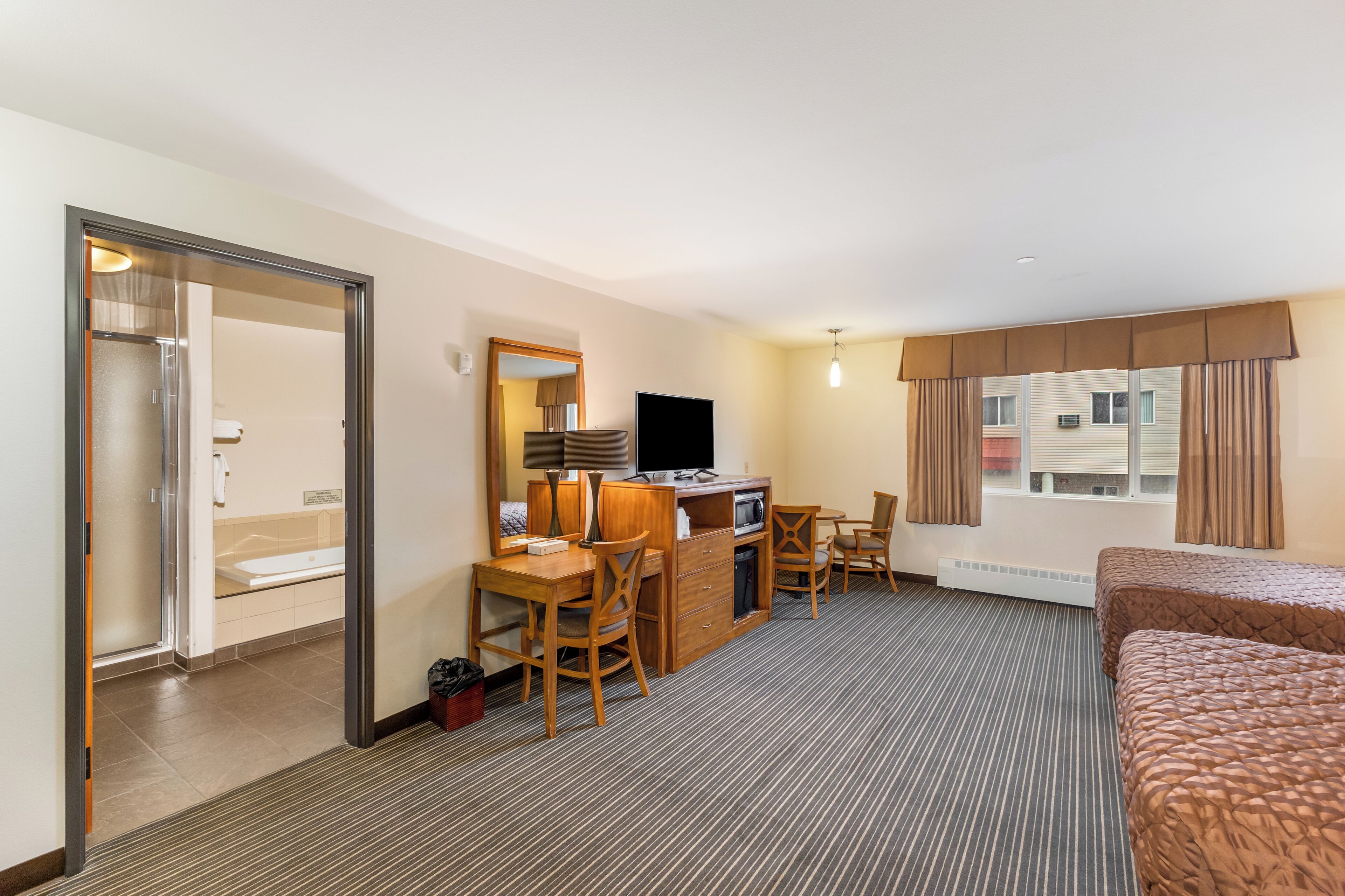 A hotel room with a bed, a desk with a chair, a television, and a bathroom visible through an open door.