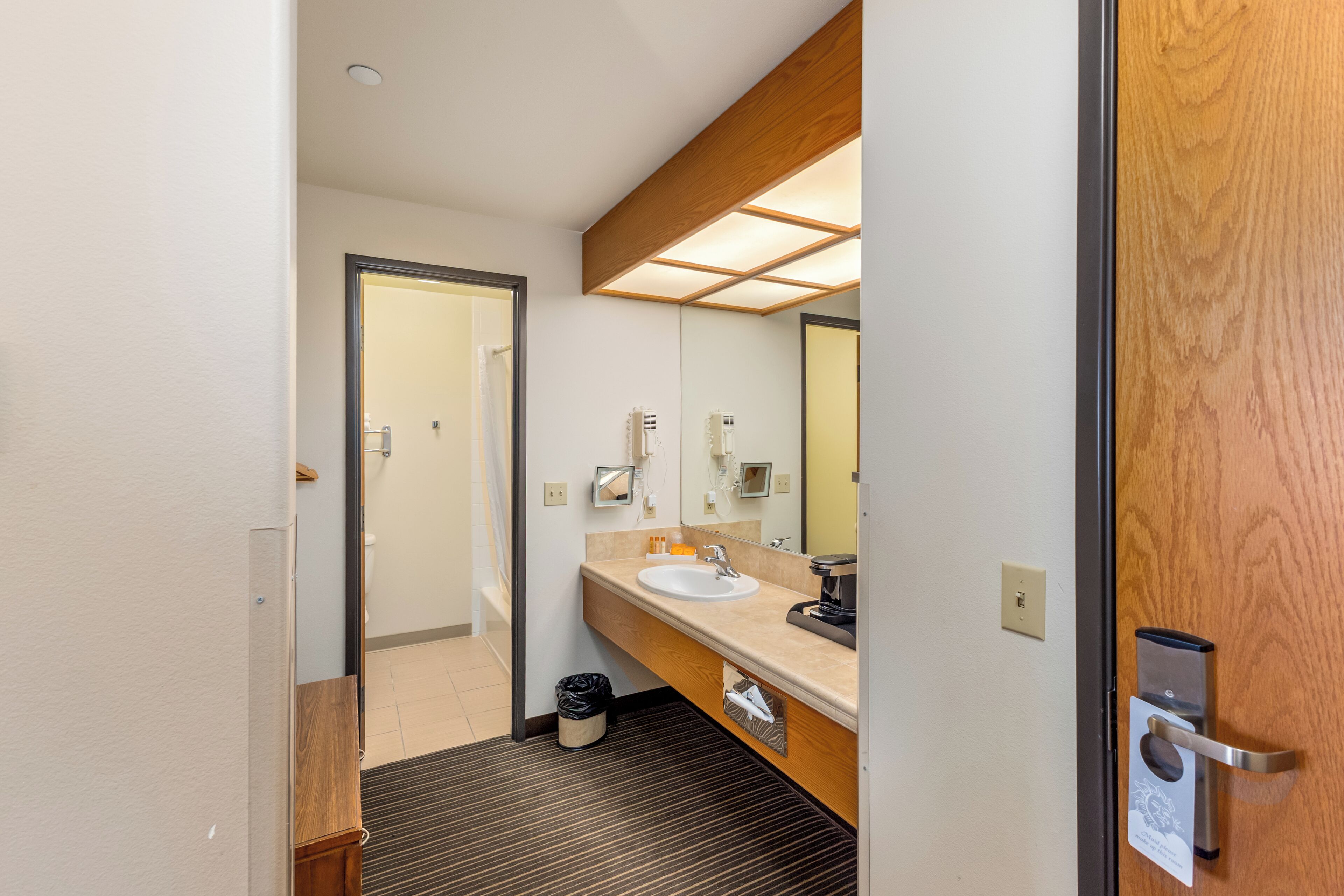 A hotel bathroom with a wooden vanity, a sink, a mirror, and a door with a handle.