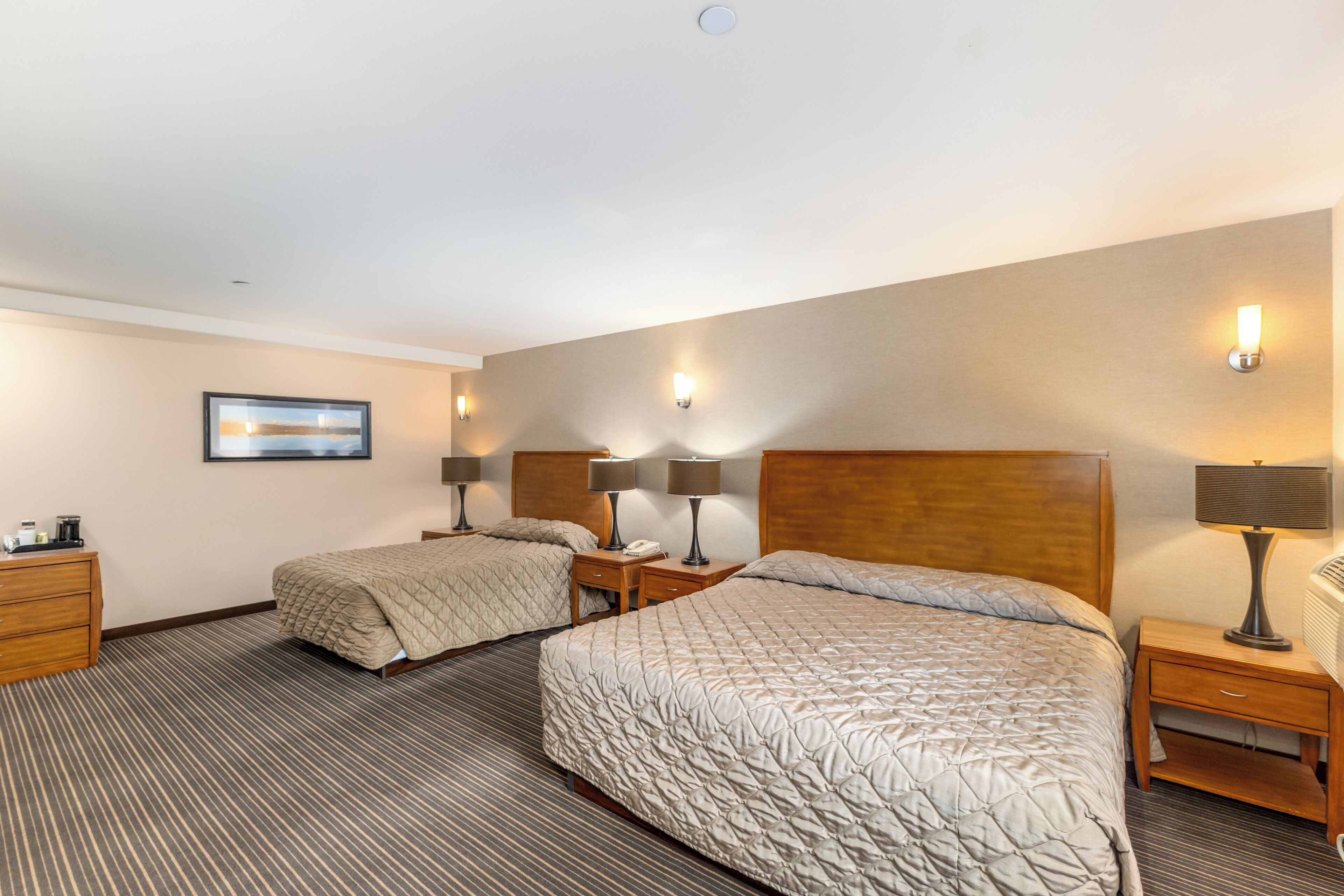 Une chambre d’hôtel avec deux lits, une tête de lit en bois, des tables de chevet avec des lampes, un tableau encadré au mur et un tapis à rayures.