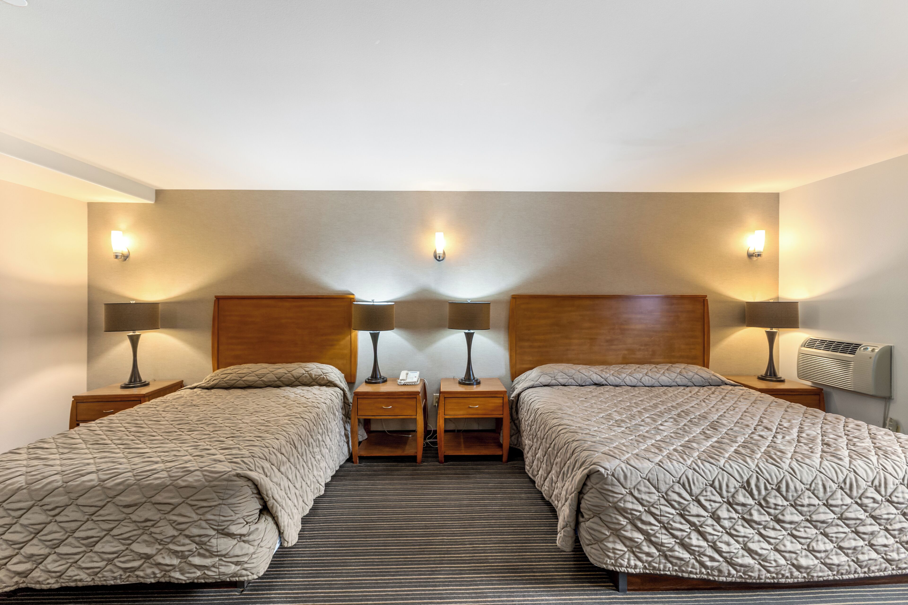 A hotel room with two beds, each with a wooden headboard, bedside tables with lamps, and a wall-mounted air conditioner.