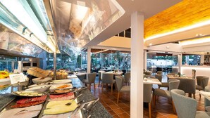 A restaurant with a buffet setup, including various meats and cheeses, and a view of the outdoors through large windows. - Bungalows Cordial Biarritz (San Bartolomé de Tirajana)