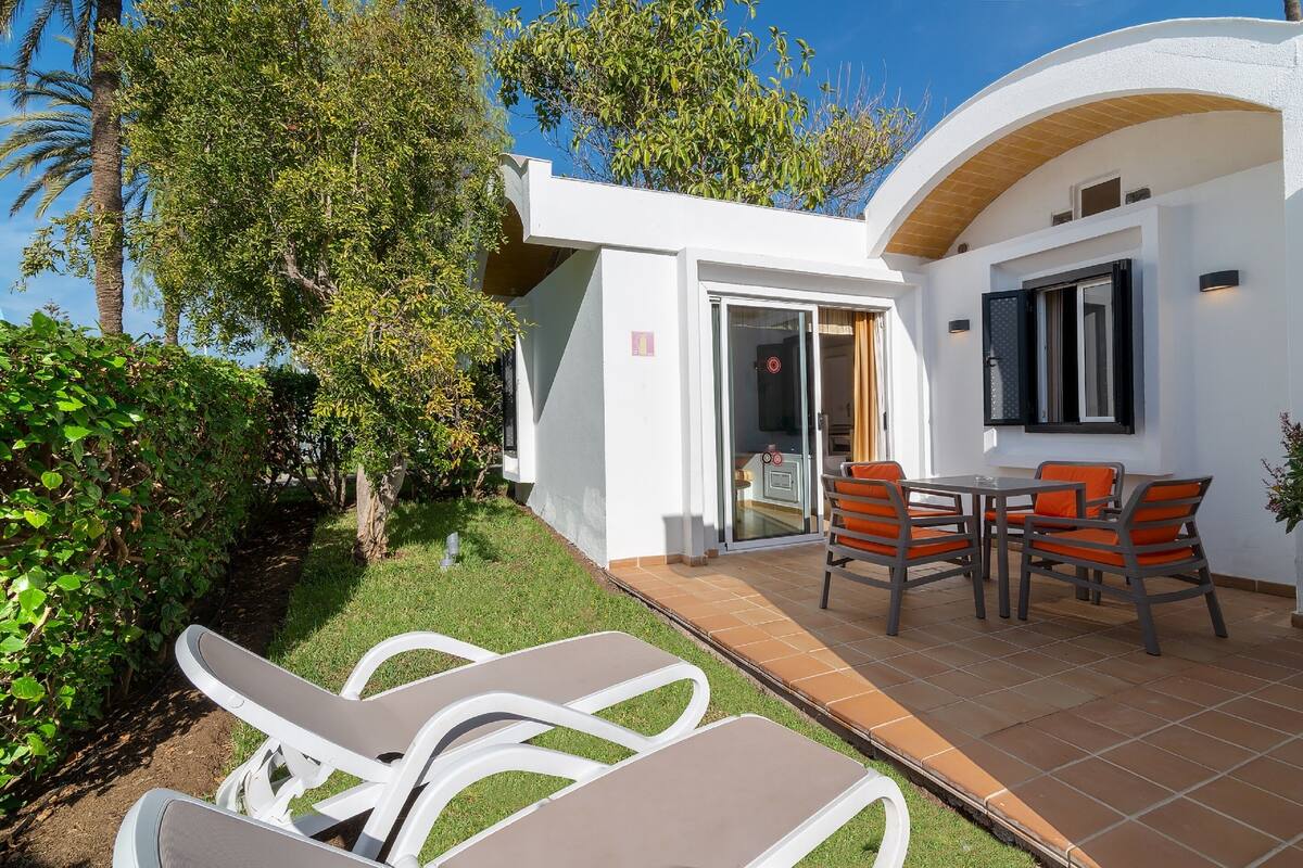 A modern house with a patio area featuring a table and chairs, surrounded by greenery and a clear sky.