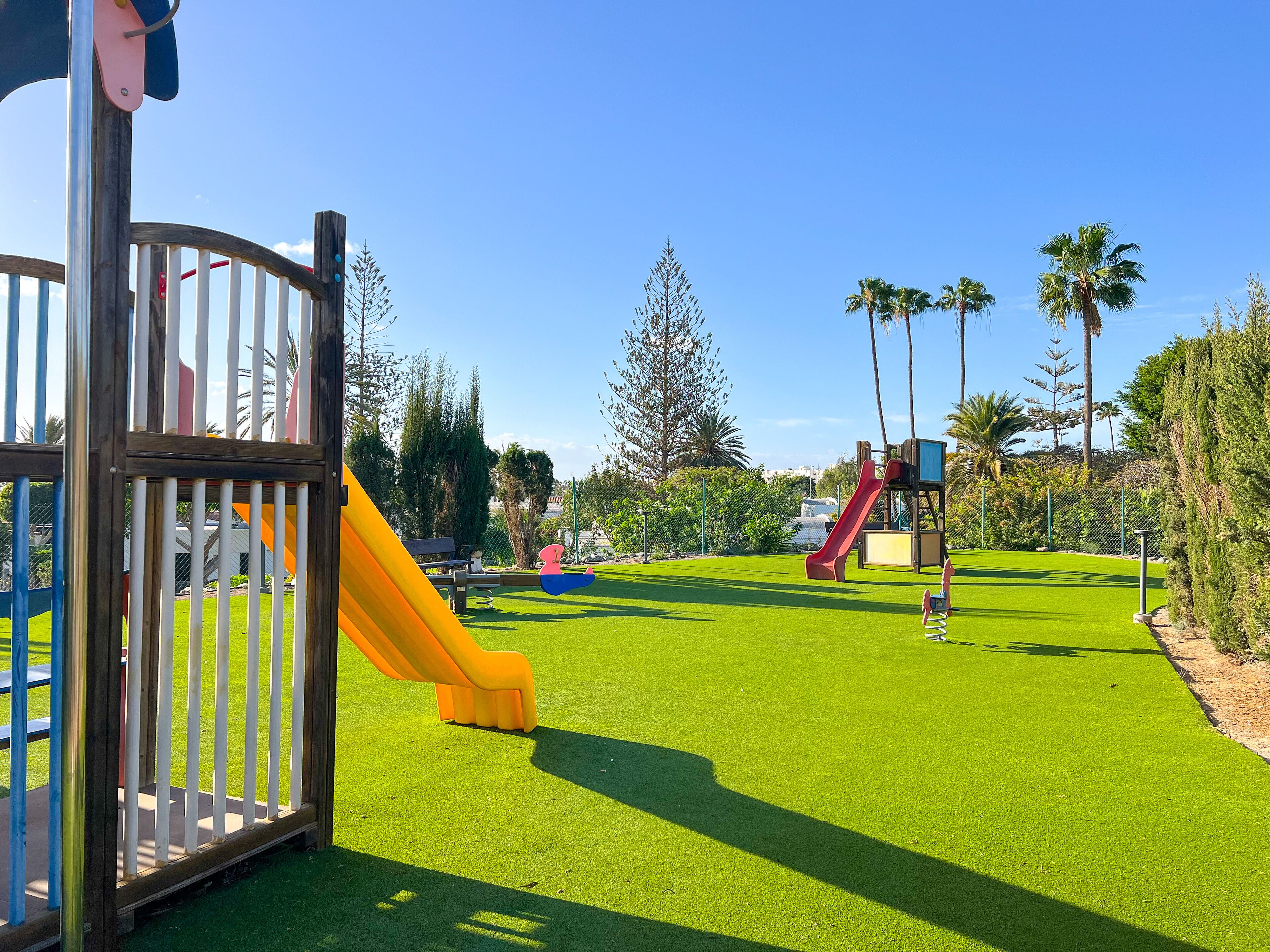 Un parque infantil con toboganes y un área cercada, rodeado de vegetación y palmeras.