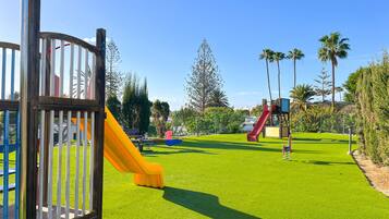 Un parque infantil con toboganes y un área cercada, rodeado de vegetación y palmeras.
