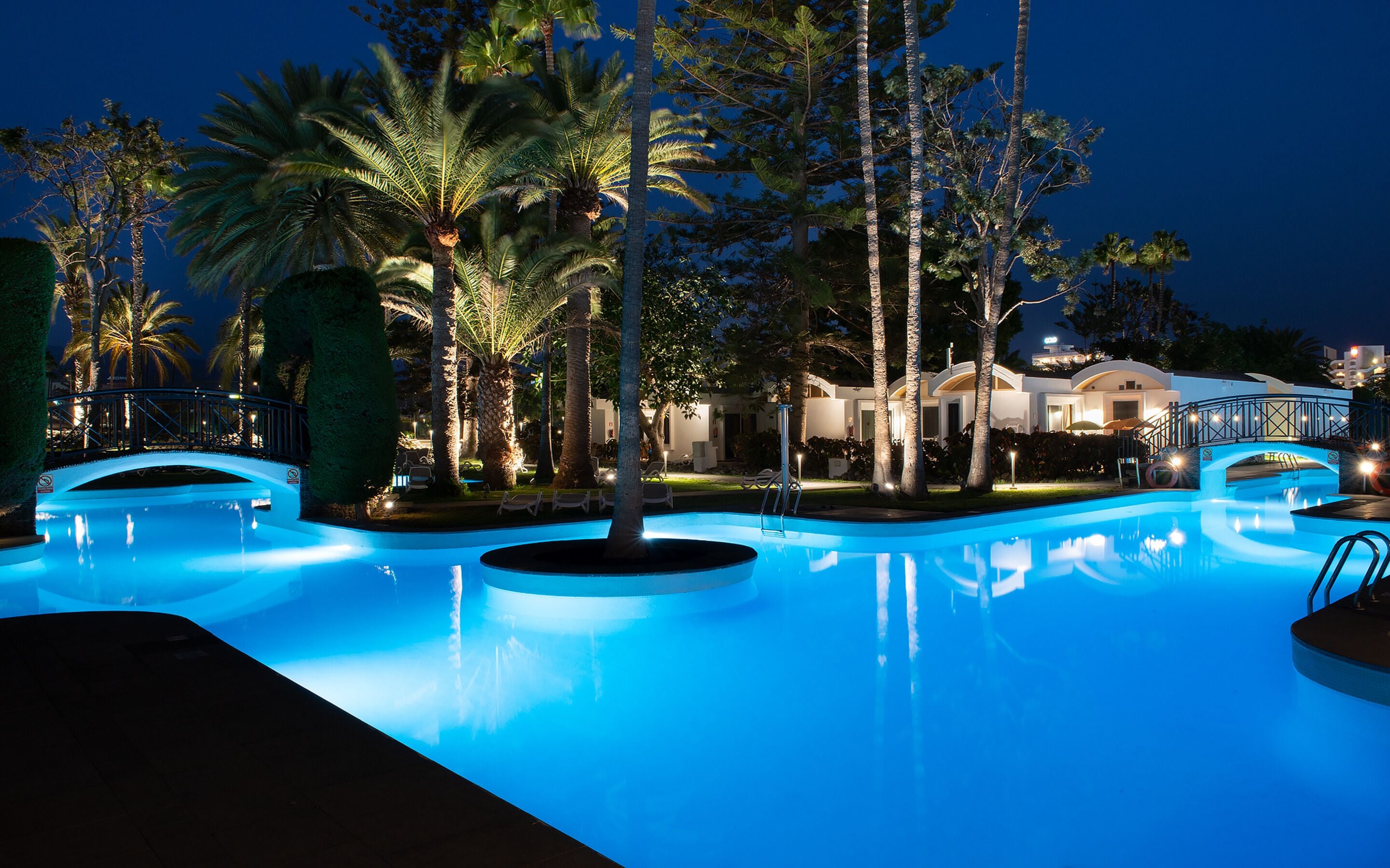 Ein beleuchteter Pool bei Nacht, umgeben von Palmen und einer Brücke.
