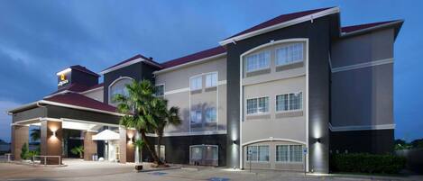 A multi-story hotel with a modern design, featuring a covered entrance, a parking lot, and a clear sky.