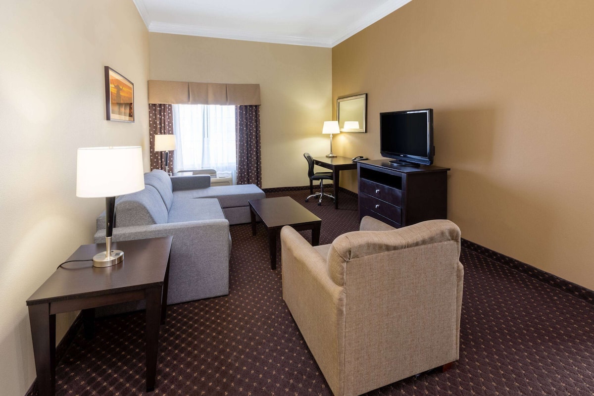 A hotel room with a sofa, armchair, desk, television, and a lamp.
