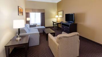 A hotel room with a sofa, armchair, desk, television, and a lamp.