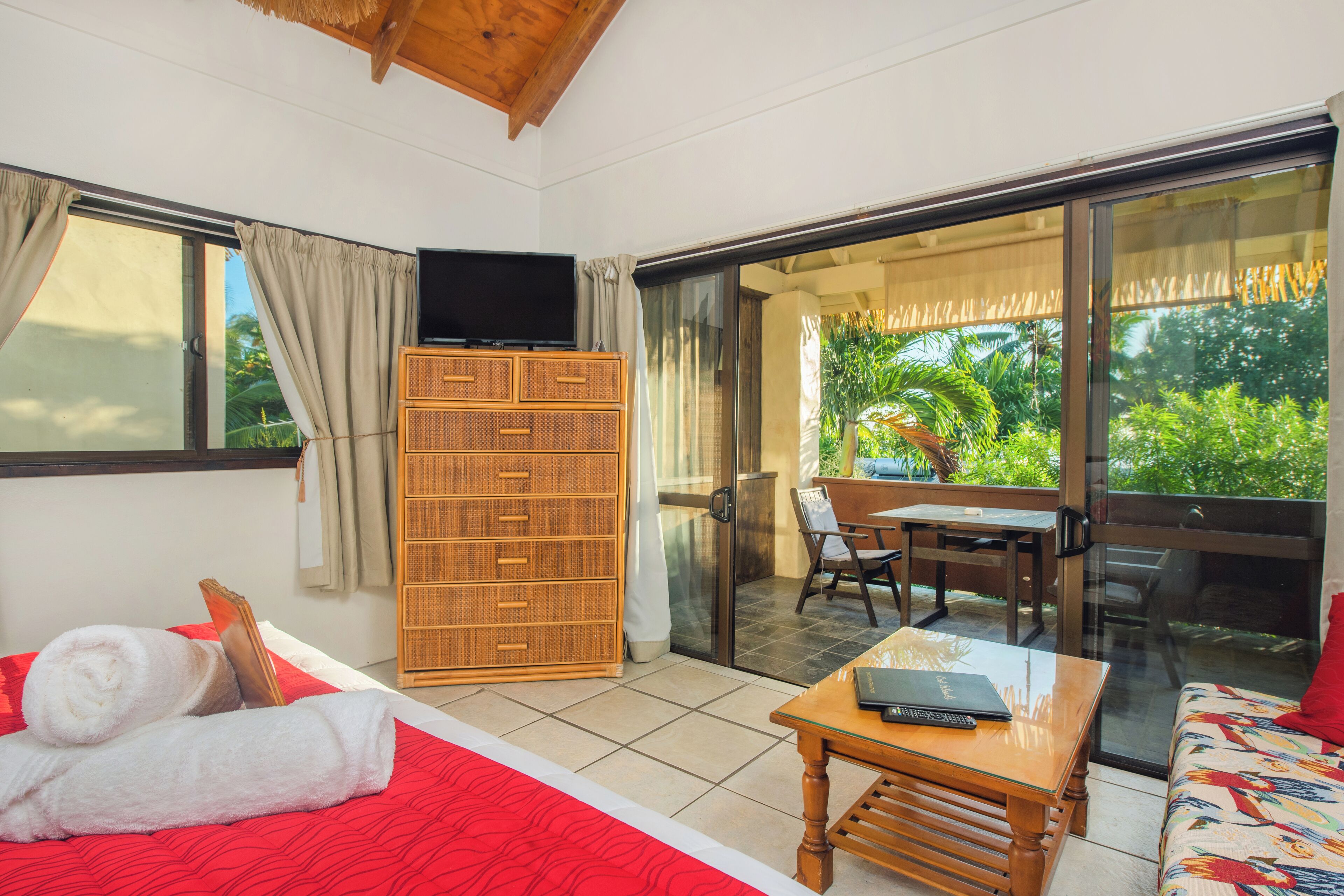 A hotel room with a bed, a wooden dresser, a table, and a balcony with a view of greenery.