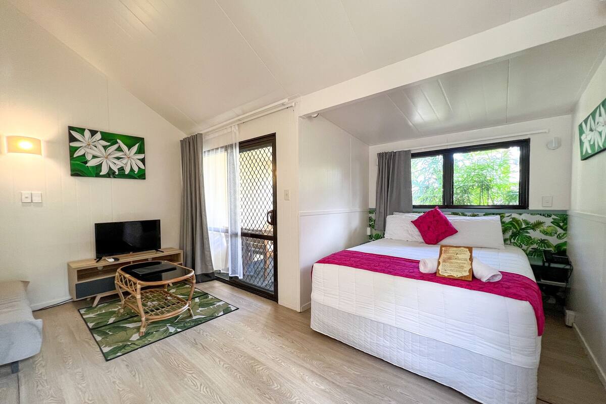 A bedroom with a bed, a TV, a small table, and a window with a view of greenery.