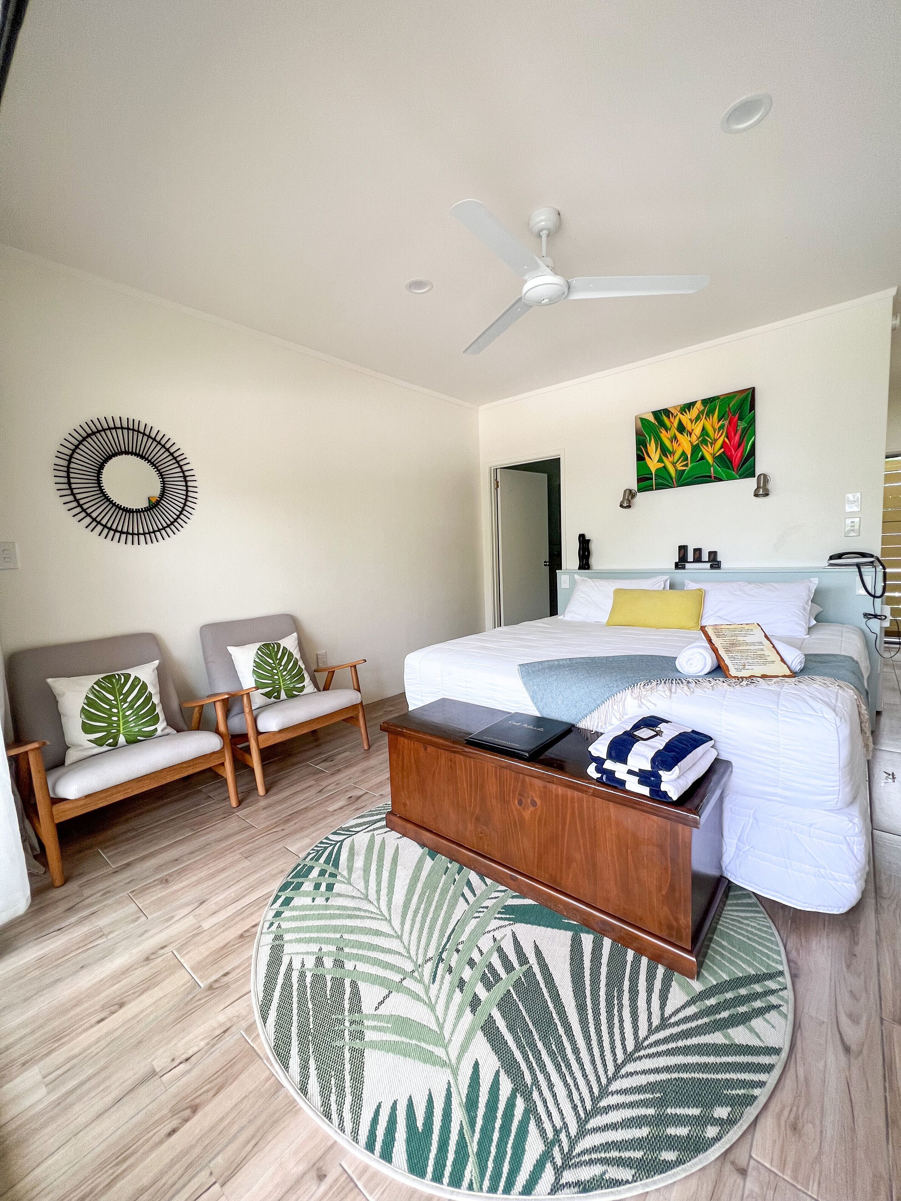 A modern bedroom with a bed, two armchairs, a coffee table, a ceiling fan, and a tropical-themed rug.