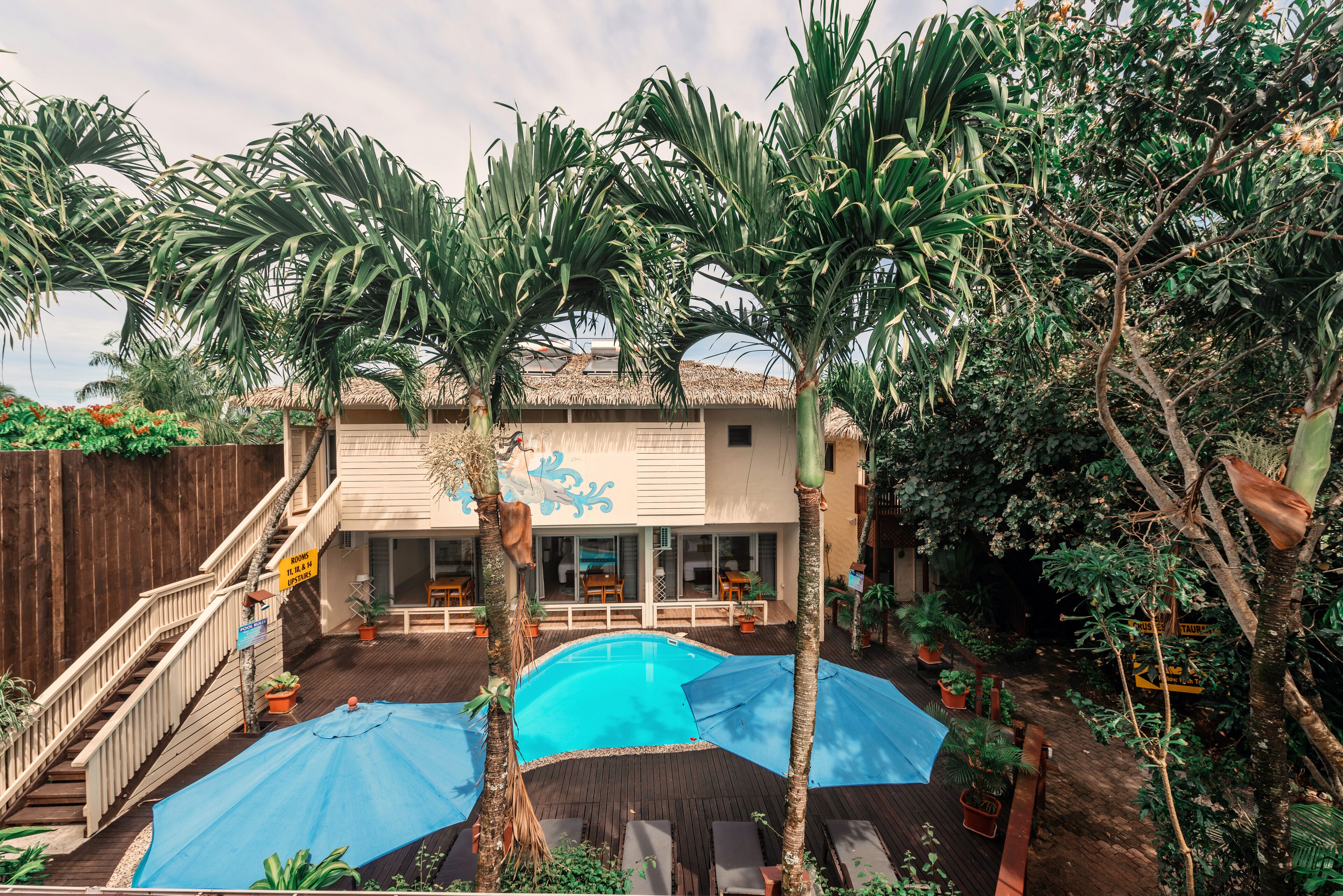 A pool area with a wooden deck, lounge chairs, and a thatched-roof building with large windows.