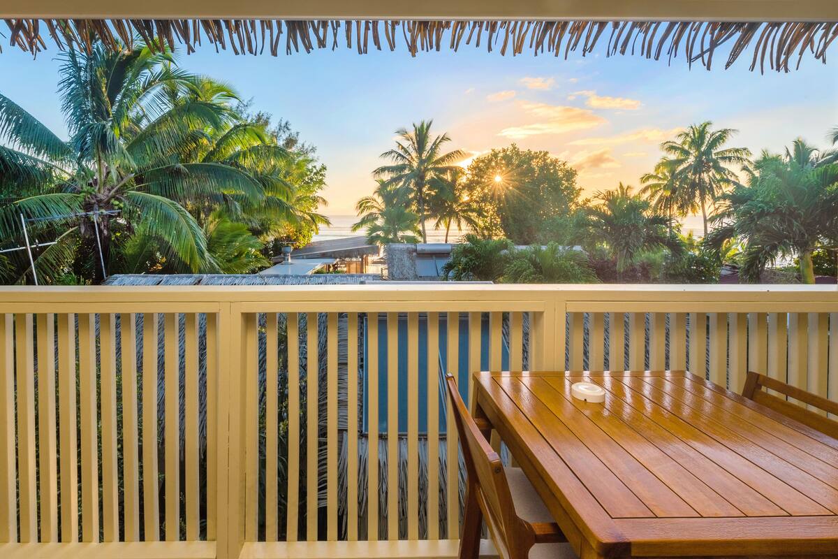 A wooden deck with a table and chairs, overlooking a beach with palm trees and a sunset.