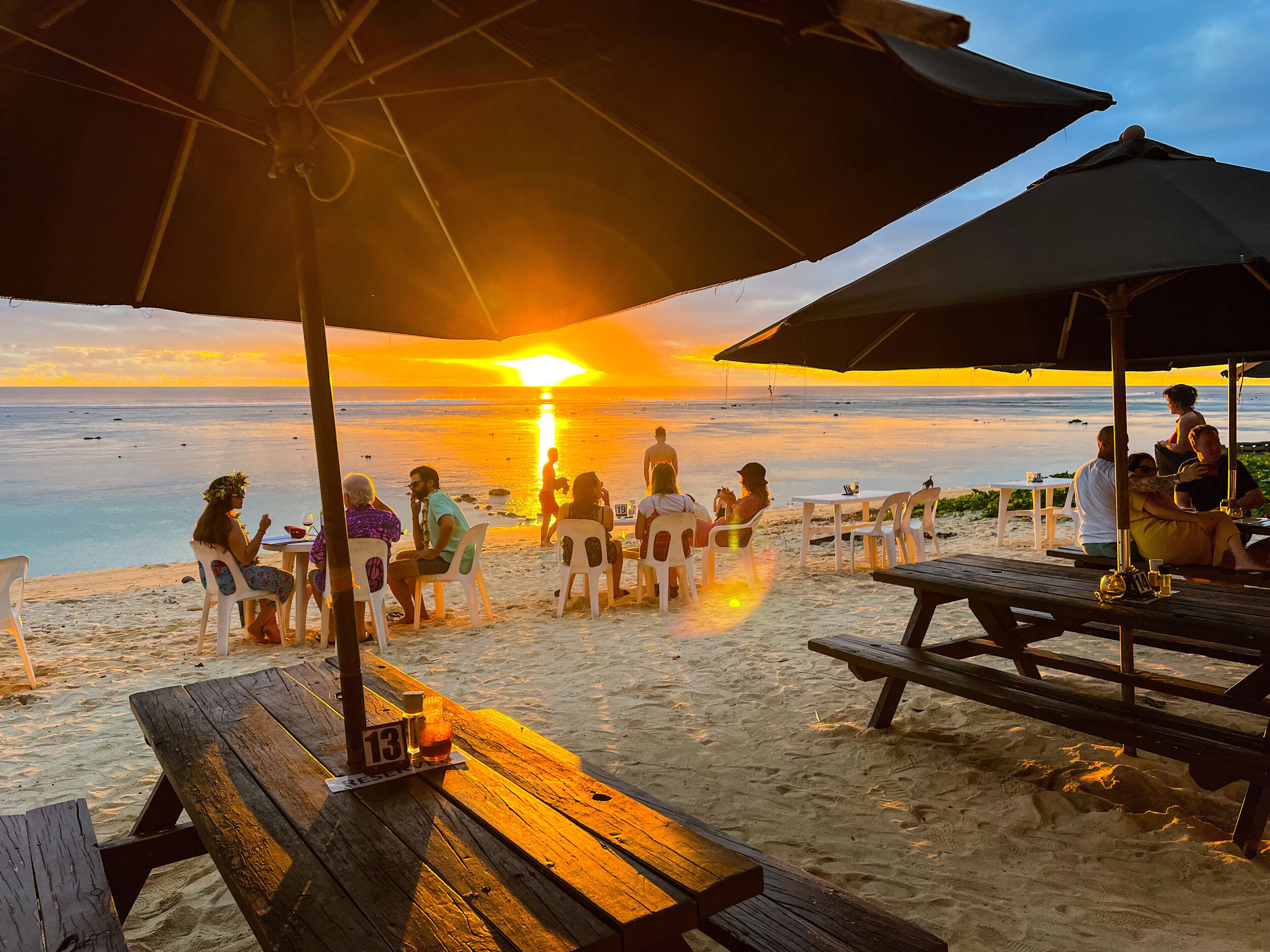 Un área de comedor frente a la playa con mesas y sillas de madera, sombrillas y vista al océano al atardecer.