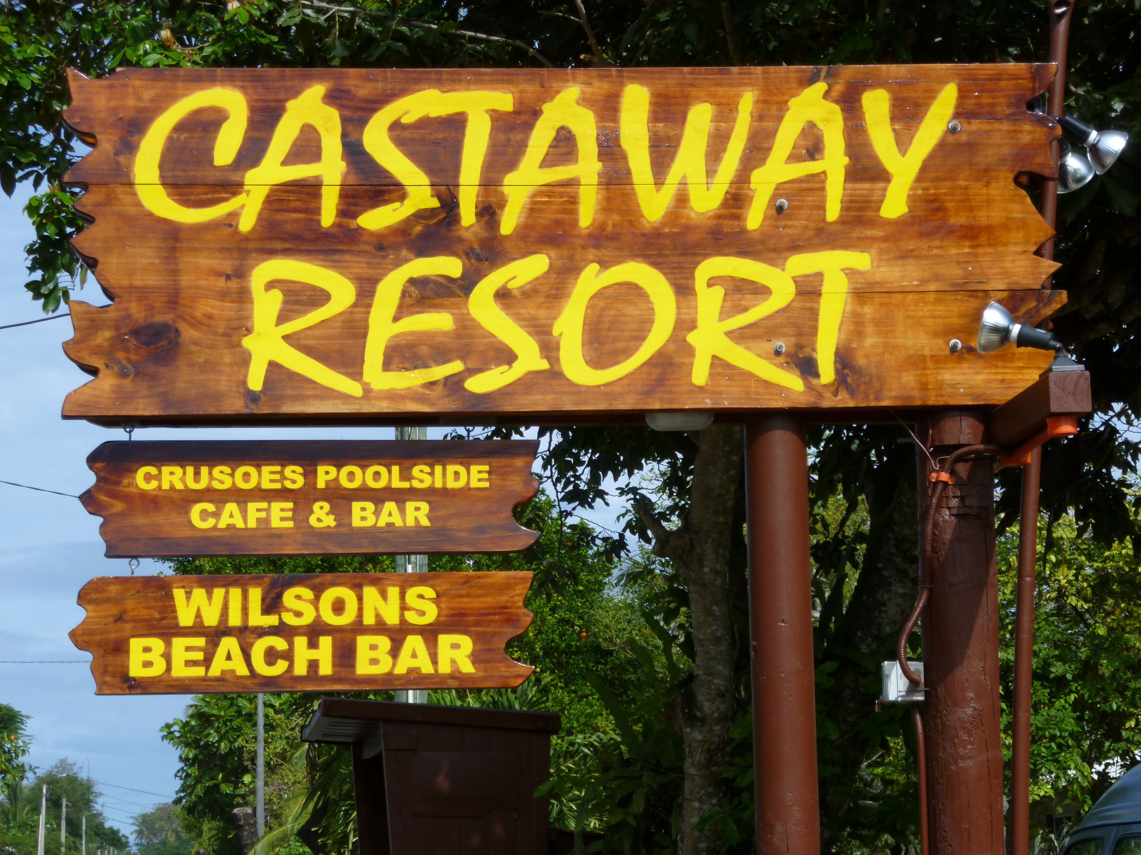 A wooden sign for Castaway Resort with yellow lettering, listing two amenities: Crusoes Poolside Cafe & Bar and Wilsons Beach Bar.