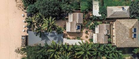 An aerial view of a coastal resort with thatched-roof cottages, palm trees, and a sandy beach.