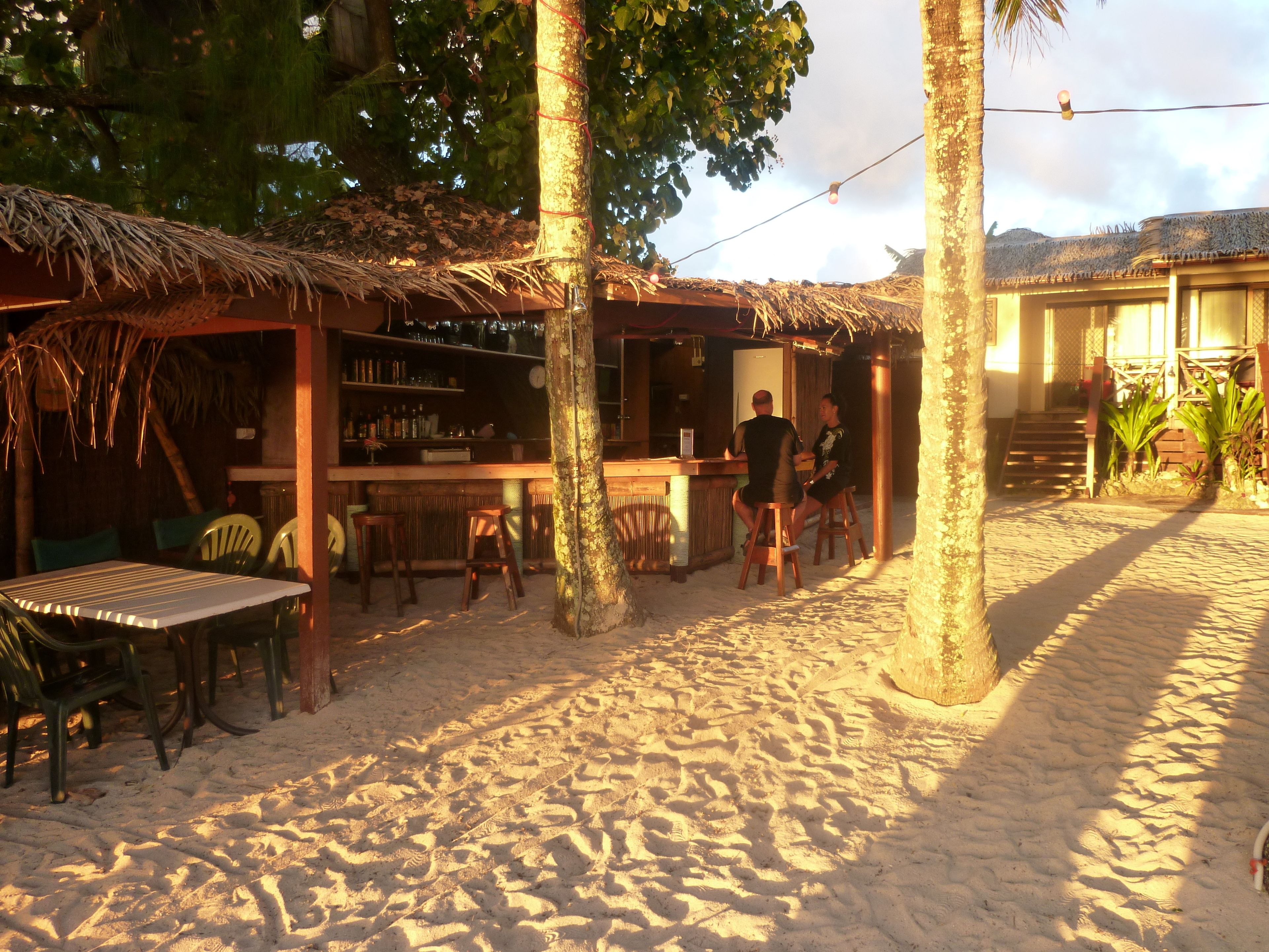 Un bar en la playa con techo de paja, mobiliario de madera y suelo de arena.