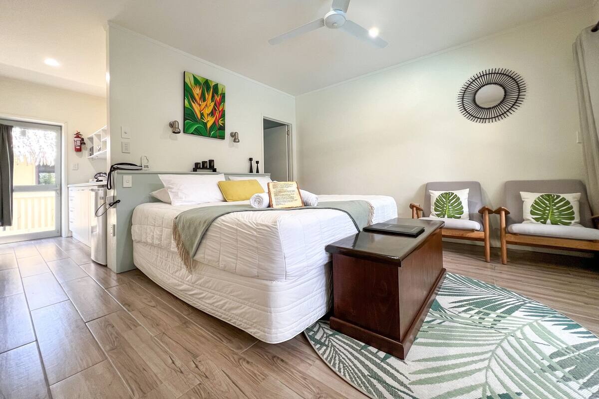 A modern bedroom with a bed, bedside tables, a desk, chairs, a ceiling fan, and a painting on the wall.