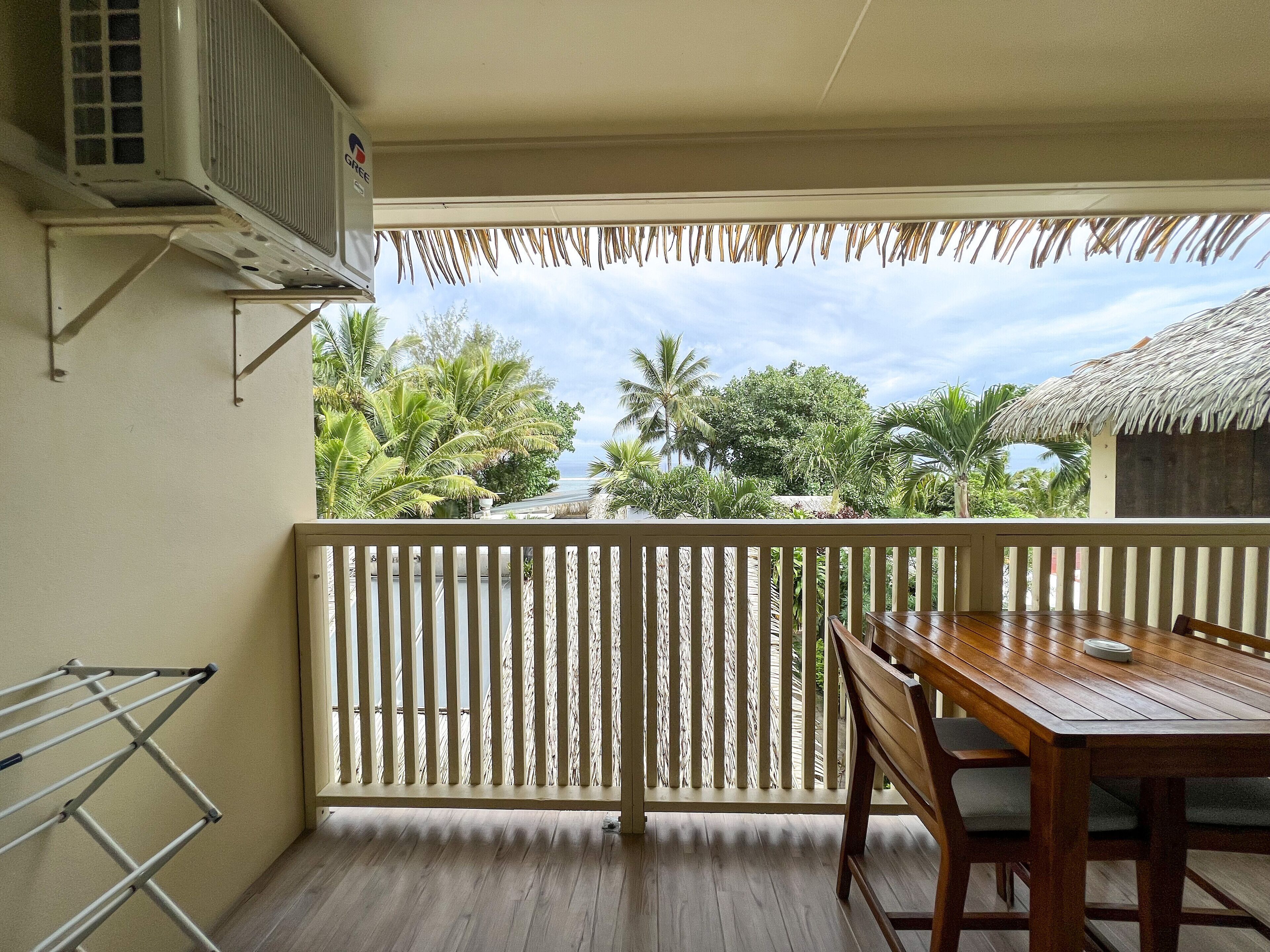 A balcony with a wooden table and chairs, a clothes drying rack, and a view of lush greenery and a thatched-roof building.
