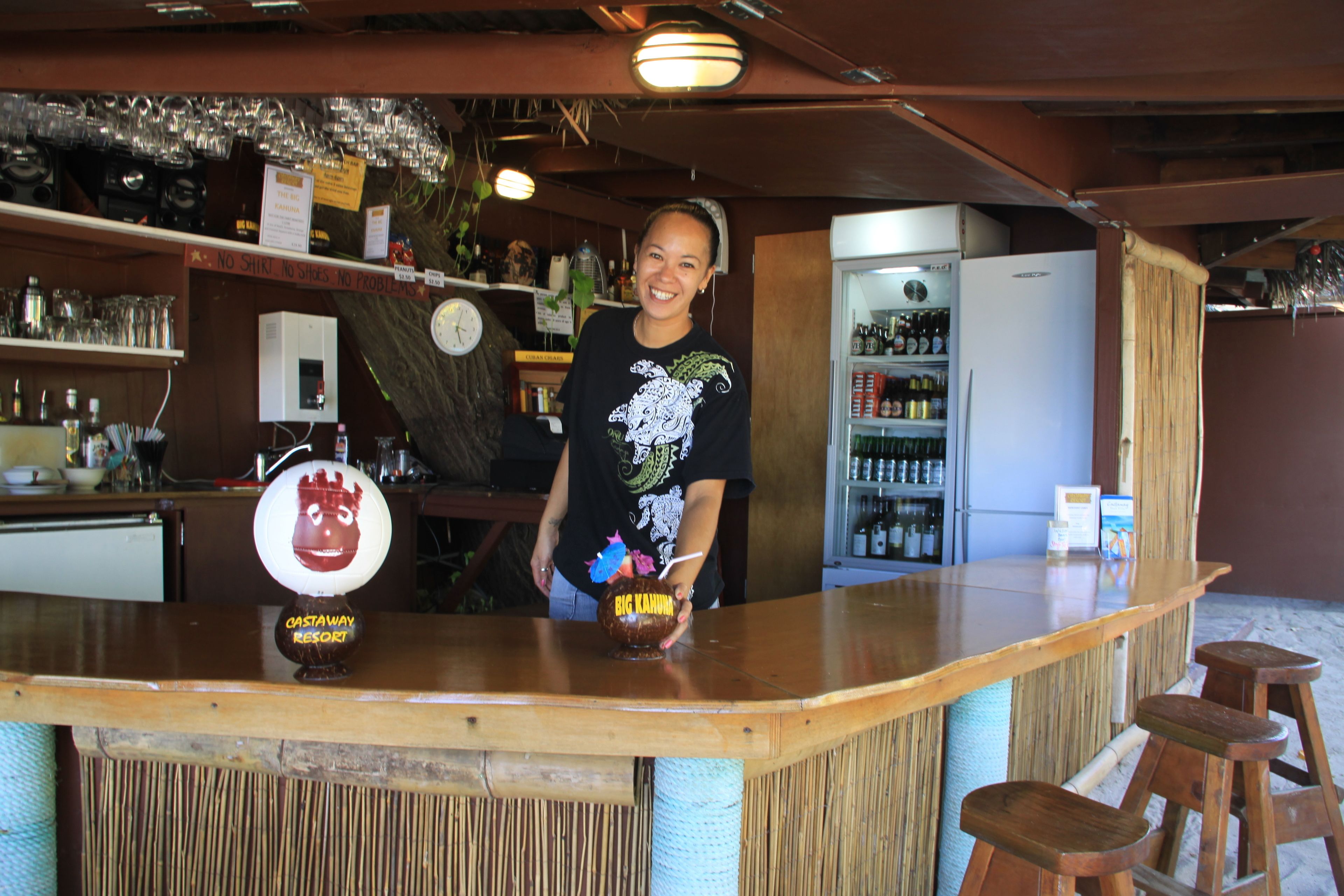 A bar area with a wooden counter, stools, and a refrigerator stocked with beverages.