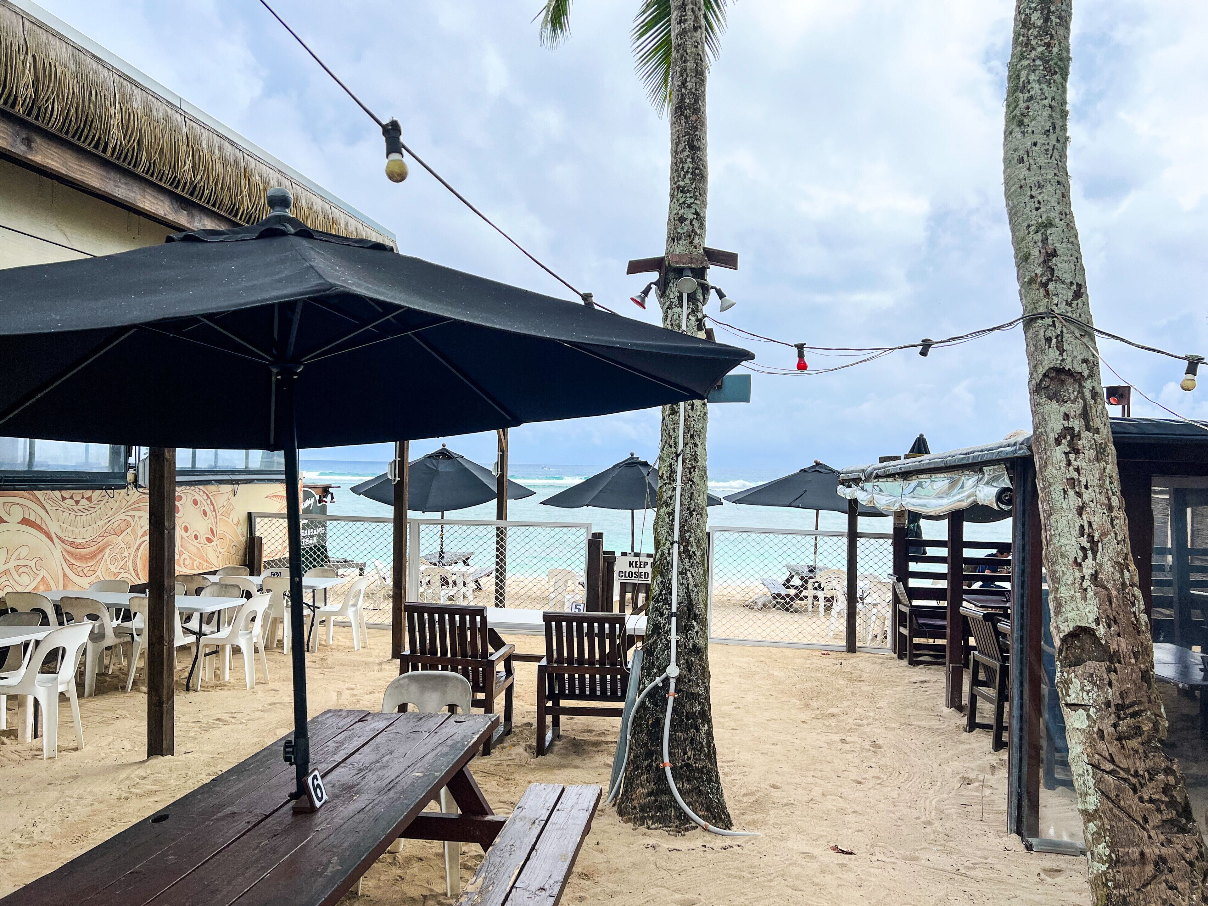 A beachside restaurant with outdoor seating, umbrellas, and a view of the ocean.
