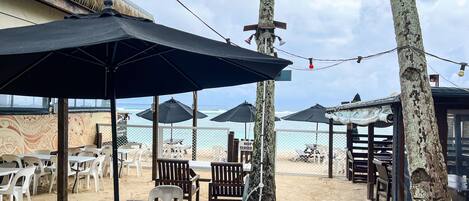 A beachside restaurant with outdoor seating, umbrellas, and a view of the ocean.