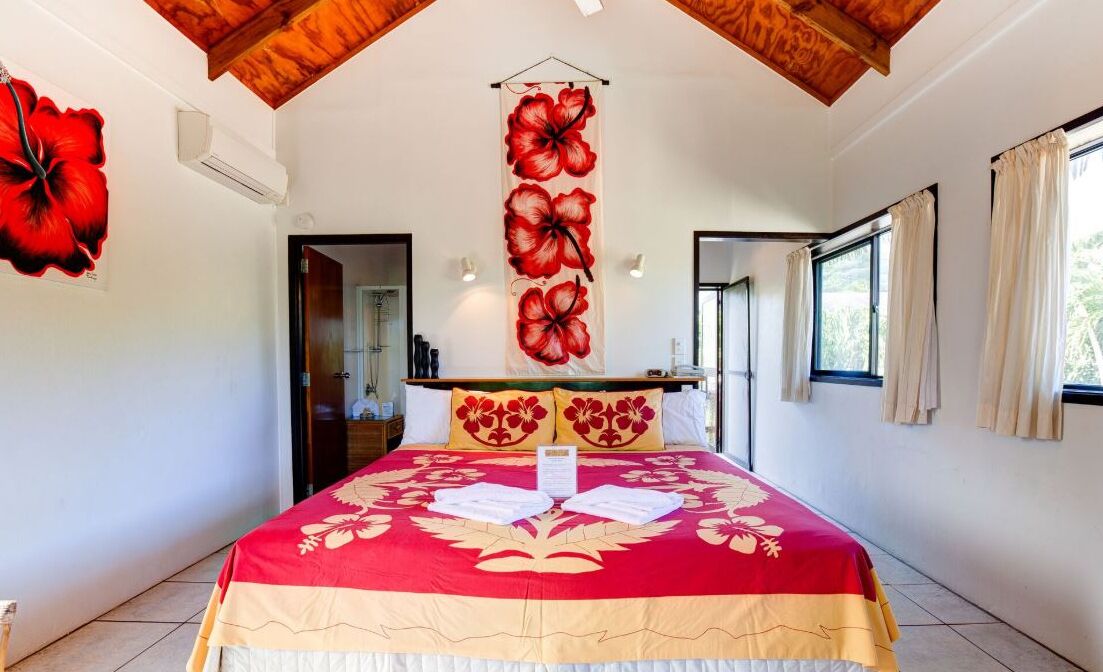 Une chambre d’hôtel avec un grand lit, un plafond en bois, une couvre-lit rouge et beige, et une décoration florale rouge sur le mur.