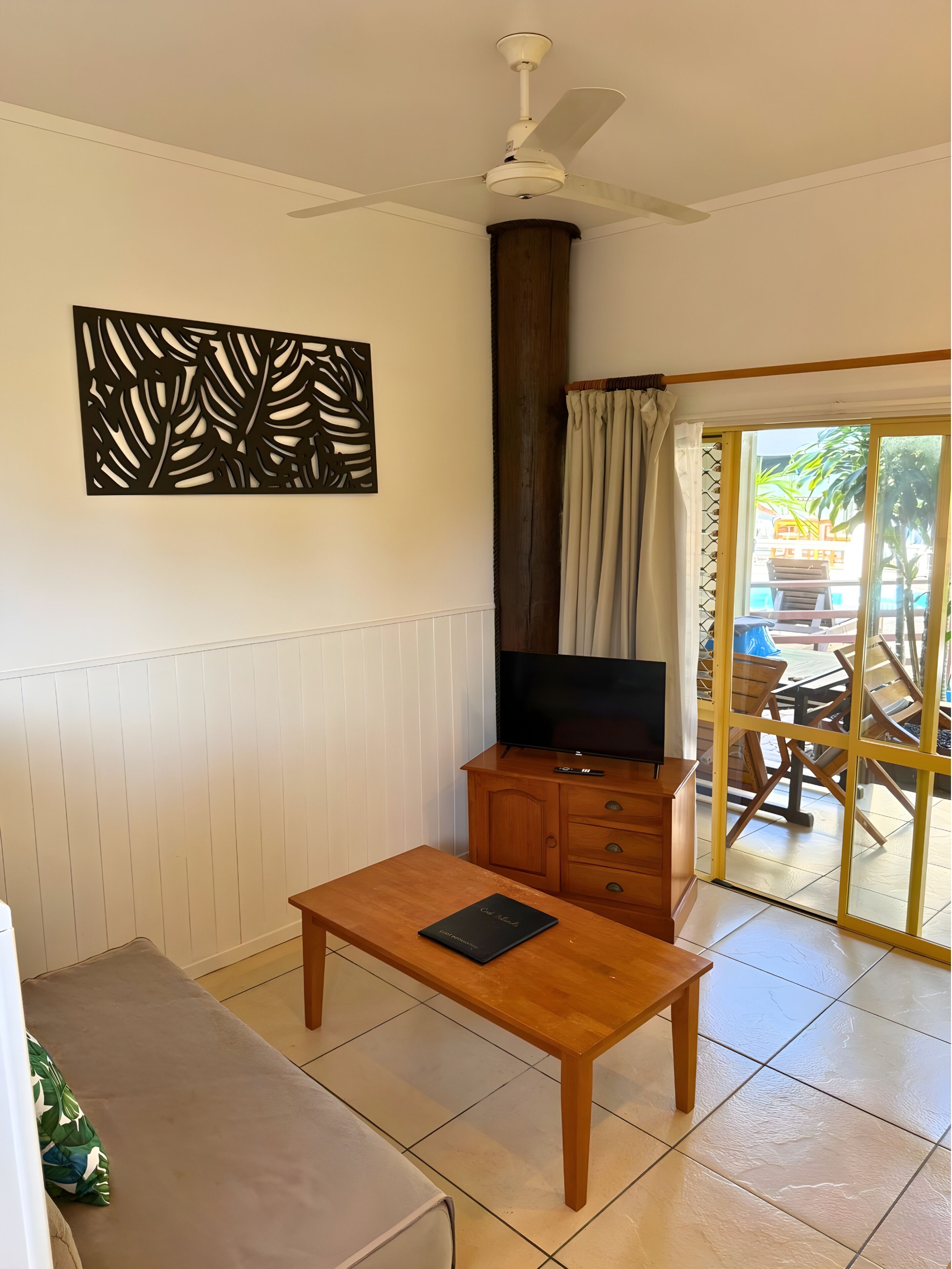 A room with a wooden table, a TV on a cabinet, a ceiling fan, a decorative wall piece, and a sliding glass door leading to an outdoor area.