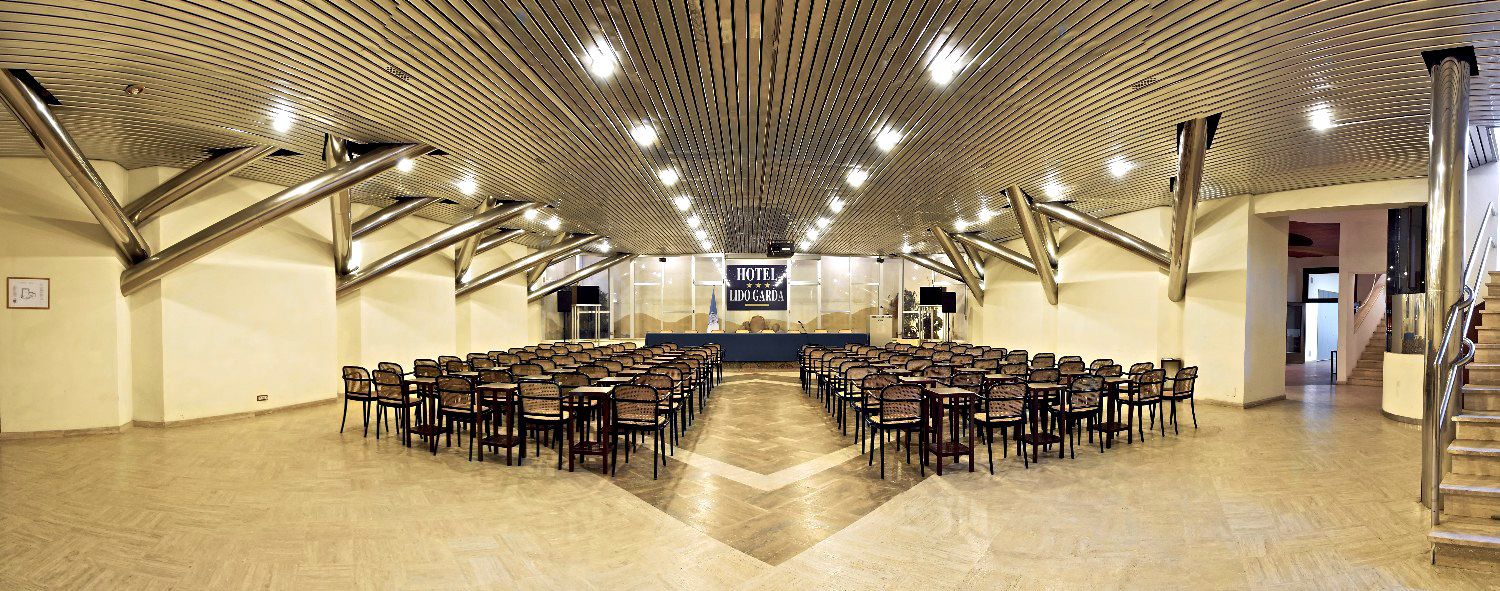 Ein geräumiger Saal mit aneinandergereihten Sitzreihen für eine Veranstaltung, eine Bühne mit einem Schild 'Hotel Lobby' und einem modernen Deckenstil.