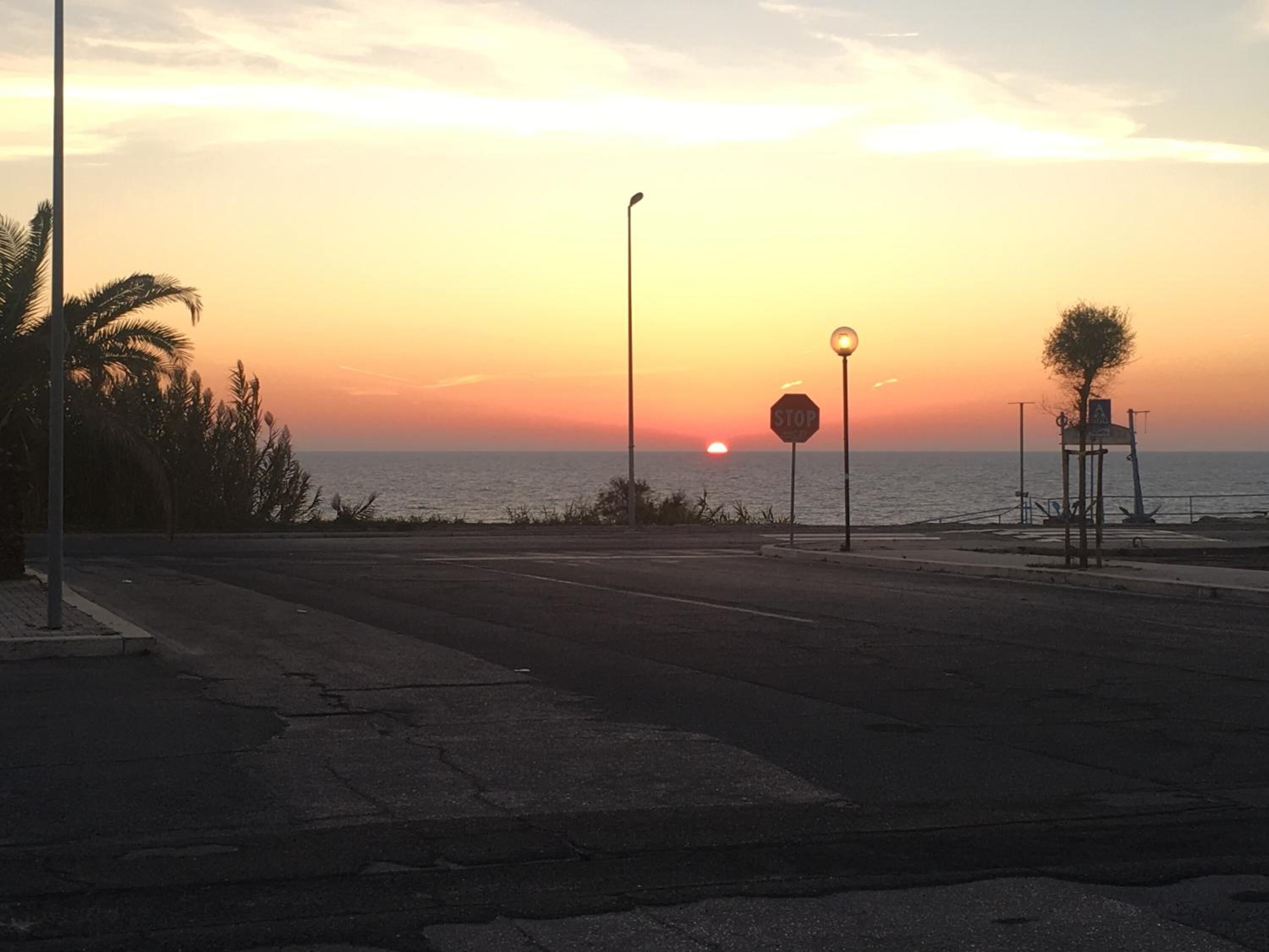 A sunset view from a coastal road with a stop sign, street lamps, and palm trees.