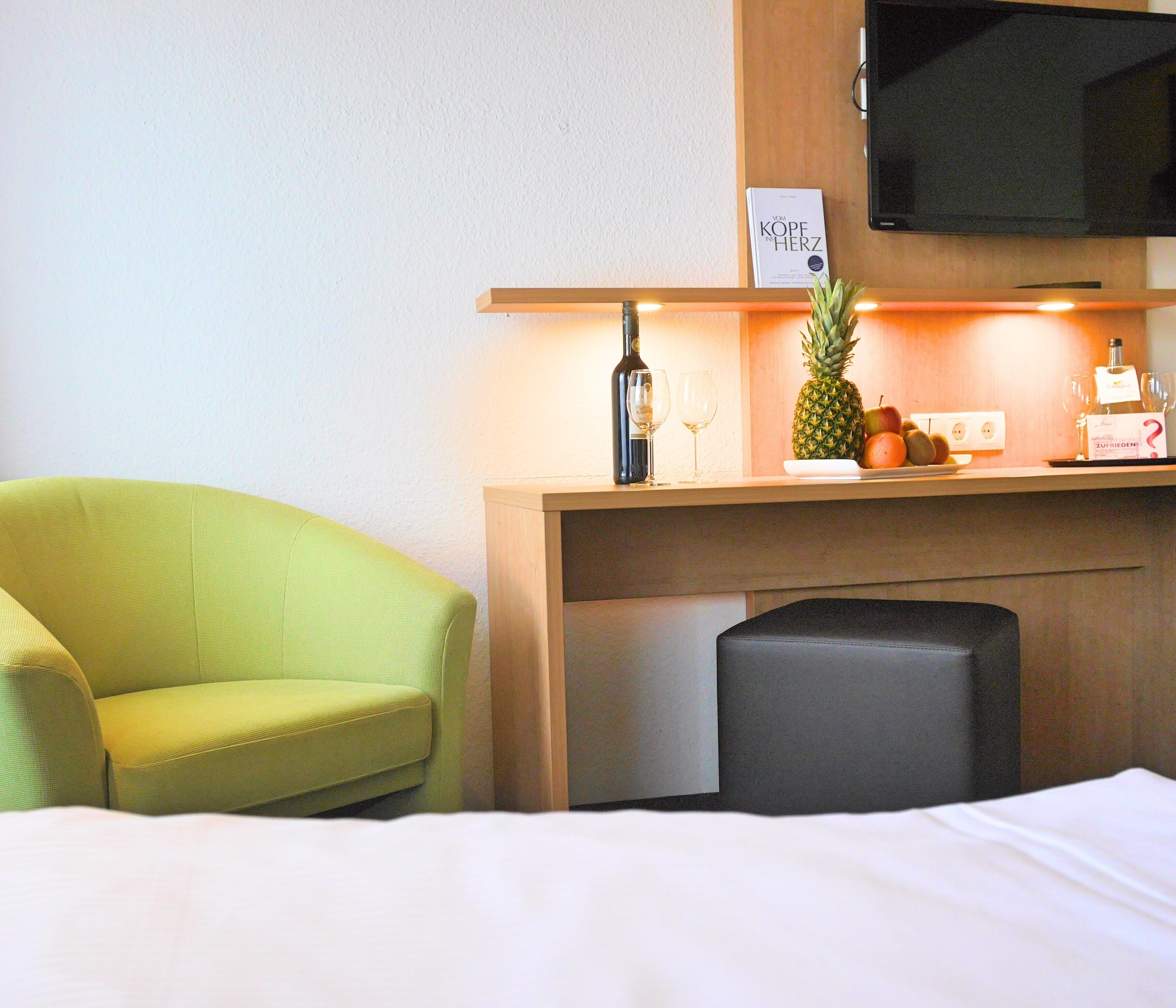 A hotel room with a bed, a green armchair, a wooden desk with a bottle of wine, a pineapple, and a fruit bowl.