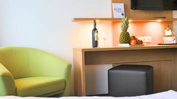 A hotel room with a bed, a green armchair, a wooden desk with a bottle of wine, a pineapple, and a fruit bowl.