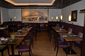Ein Restaurant mit Holztischen und violetten Sitzbänken, dessen Rückwand ein Wandbild eines Gebäudes zeigt.