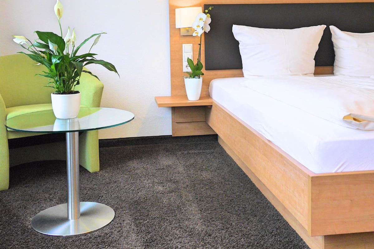 A hotel room with a bed, a green chair, a small table with a potted plant, and a nightstand with a lamp.