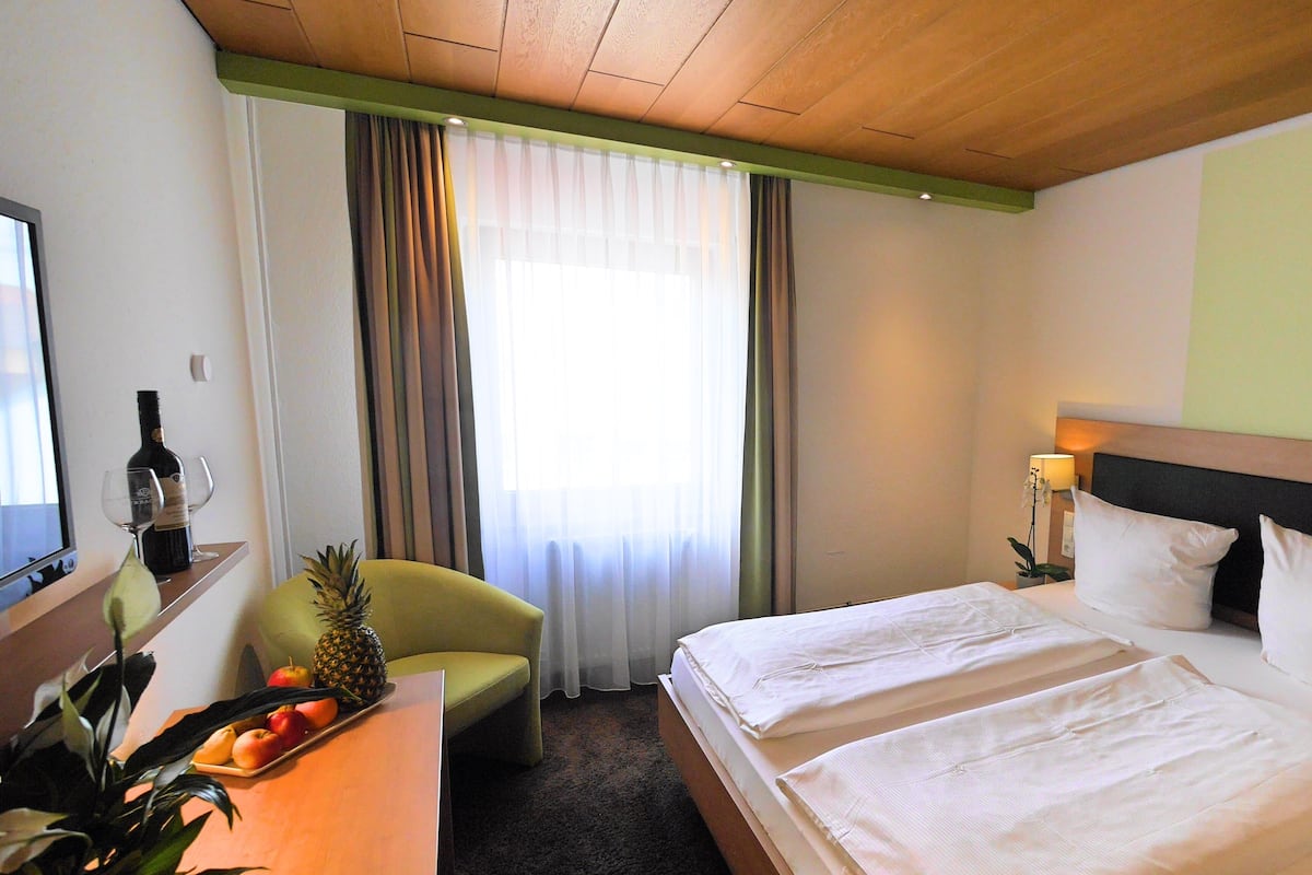 A hotel room with a bed, a desk with a bottle and glasses, a chair, a plant, and a window with curtains.
