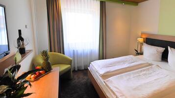 A hotel room with a bed, a desk with a bottle and glasses, a chair, a plant, and a window with curtains.