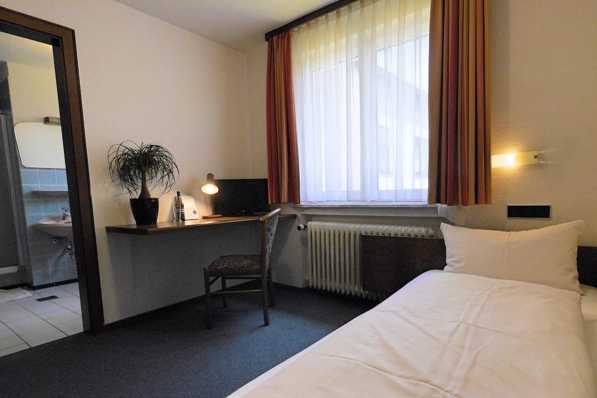 A hotel room with a bed, a desk with a computer, a chair, a potted plant, and a window with curtains.