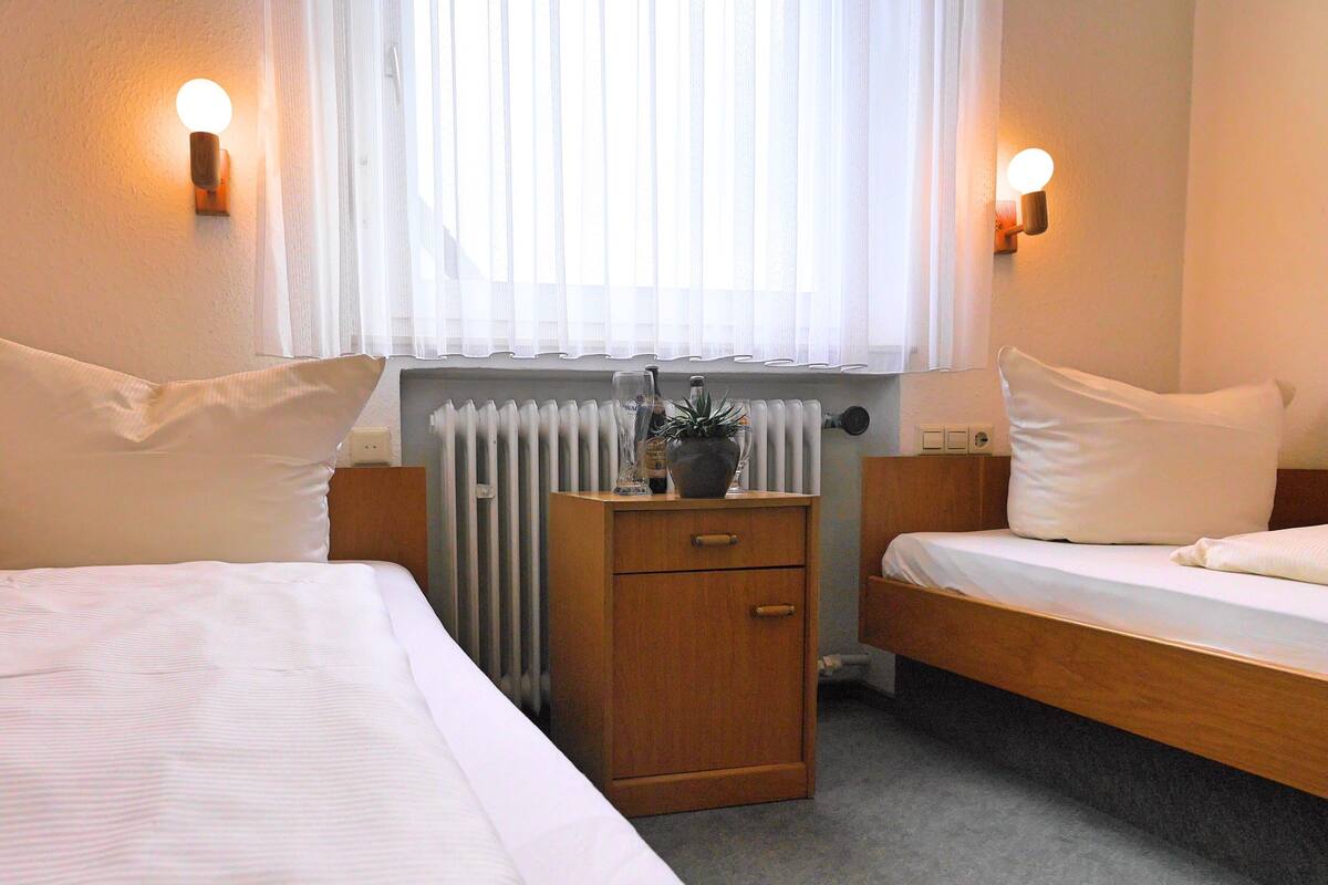 A hotel room with two beds, a wooden nightstand, a radiator, and wall-mounted lights.
