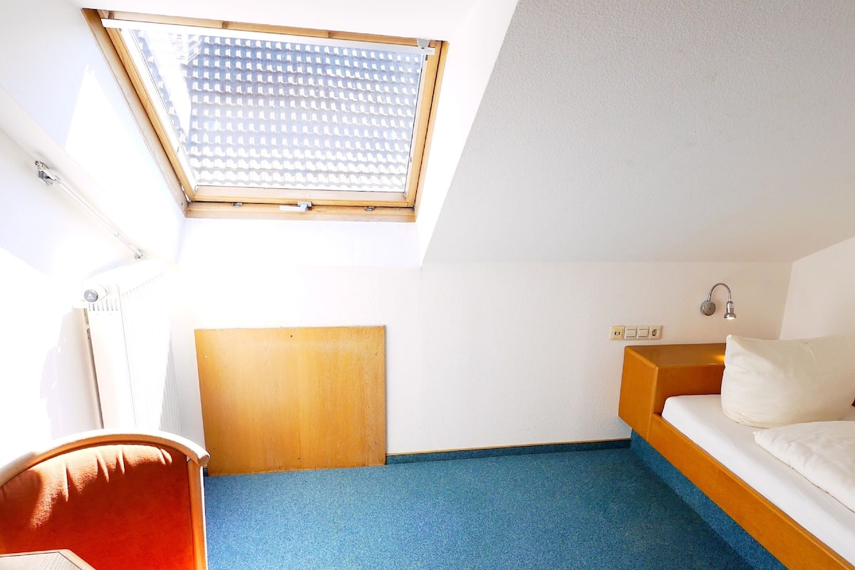 A small, bright room with a single bed, a desk, and a window with blinds.