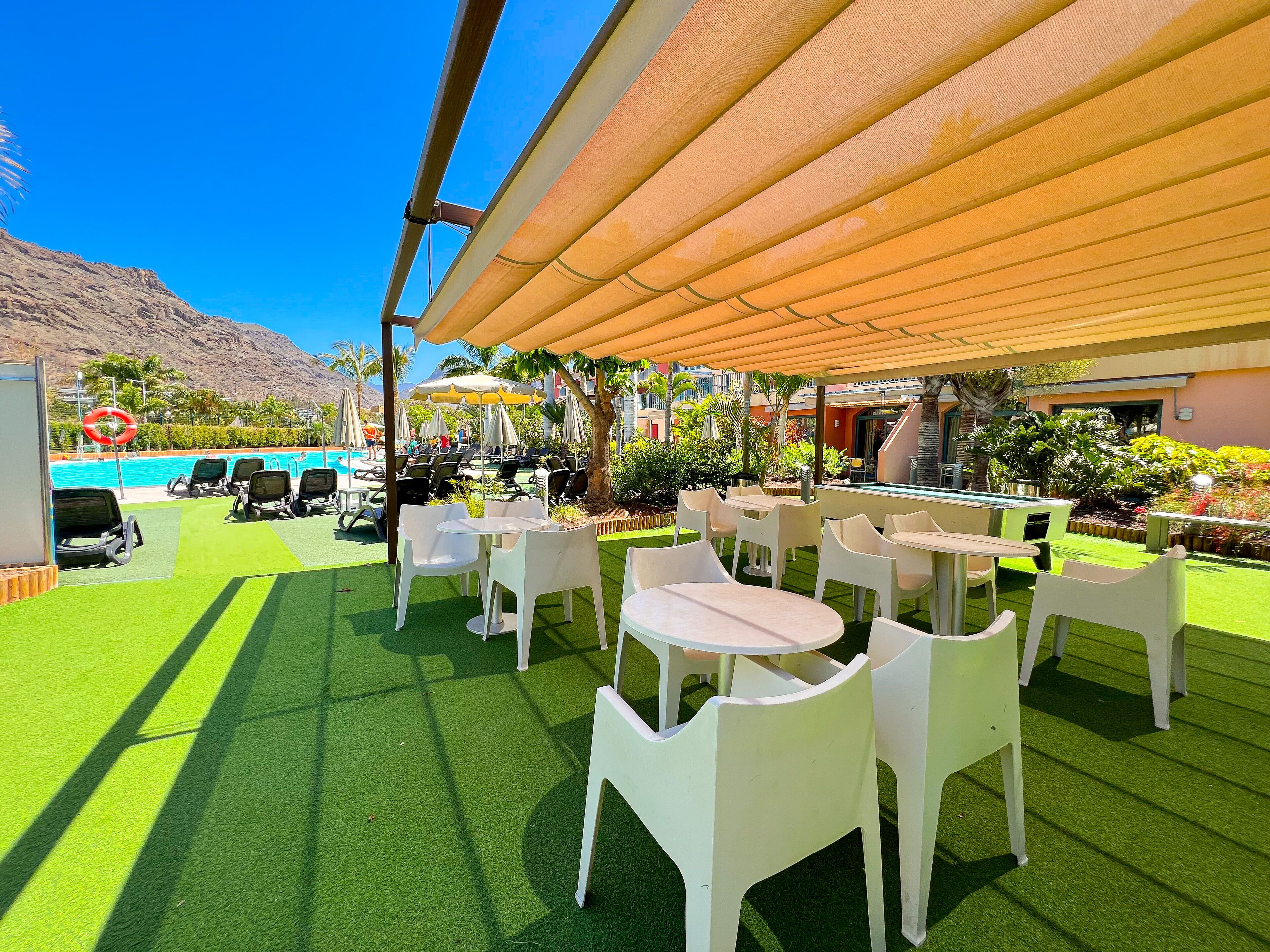Een terras buiten met witte plastic stoelen en tafels, een zwembad en parasols.