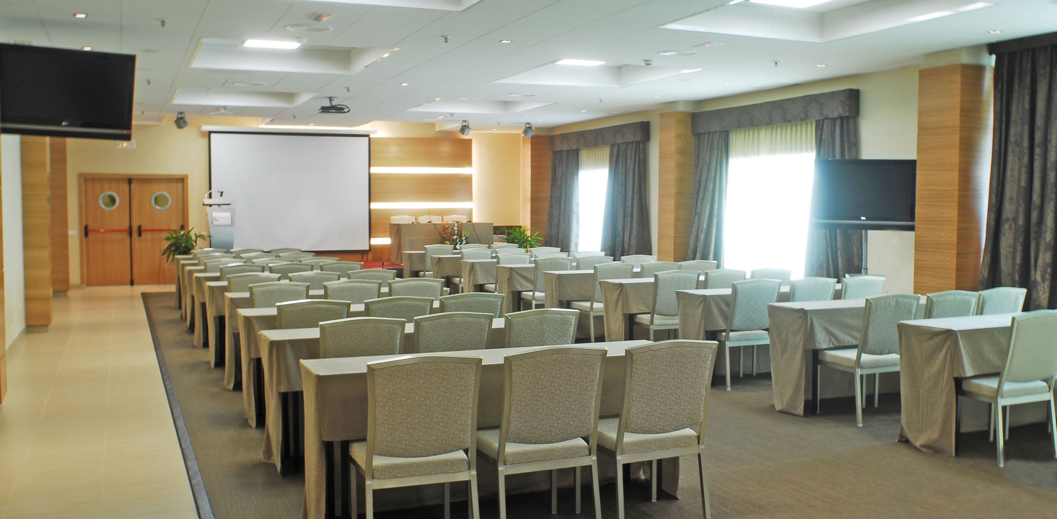A conference room with rows of tables and chairs, a projector screen, and a large TV screen.