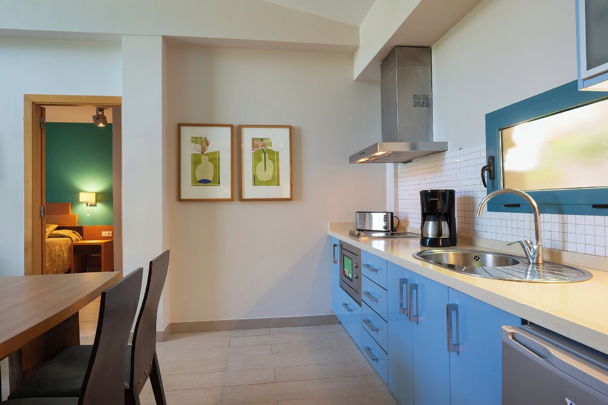 Une cuisine compacte équipée d’un évier, d’une cuisinière et d’un four à micro-ondes. Des tableaux encadrés ornent le mur, et un coin repas comprend une table et des chaises.