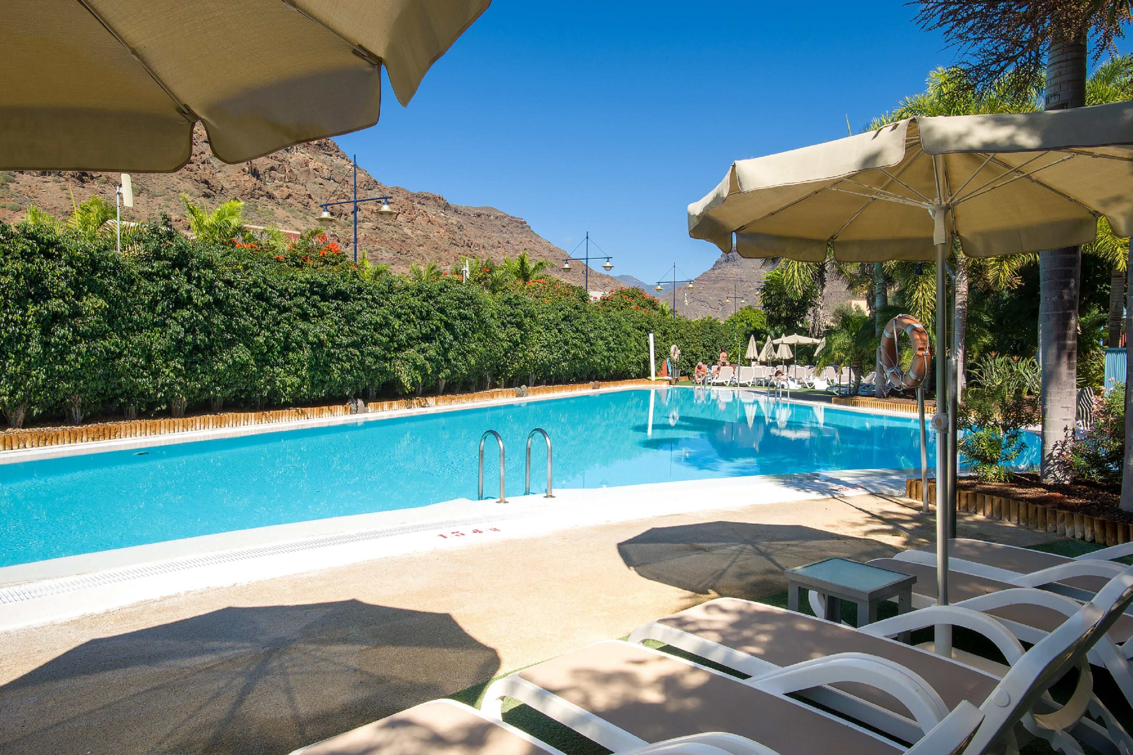 A swimming pool with lounge chairs and umbrellas, surrounded by greenery and mountains.