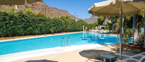 Piscina com espreguiçadeiras e guarda-sóis, cercada por vegetação e montanhas.